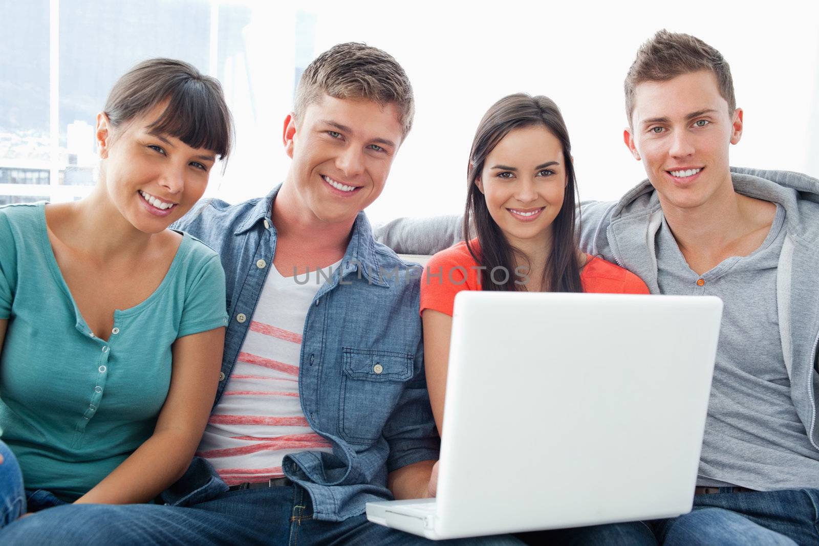 A group of friends sit on the couch together as they hold a laptop and look a the camera 