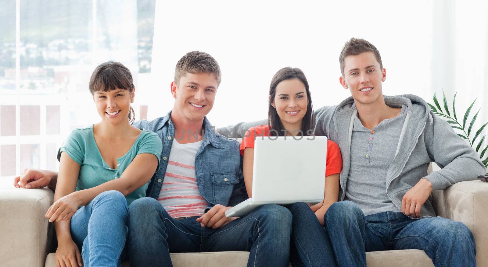 A smiling group sit on the couch as they look at the camera while they hold a laptop in front of them