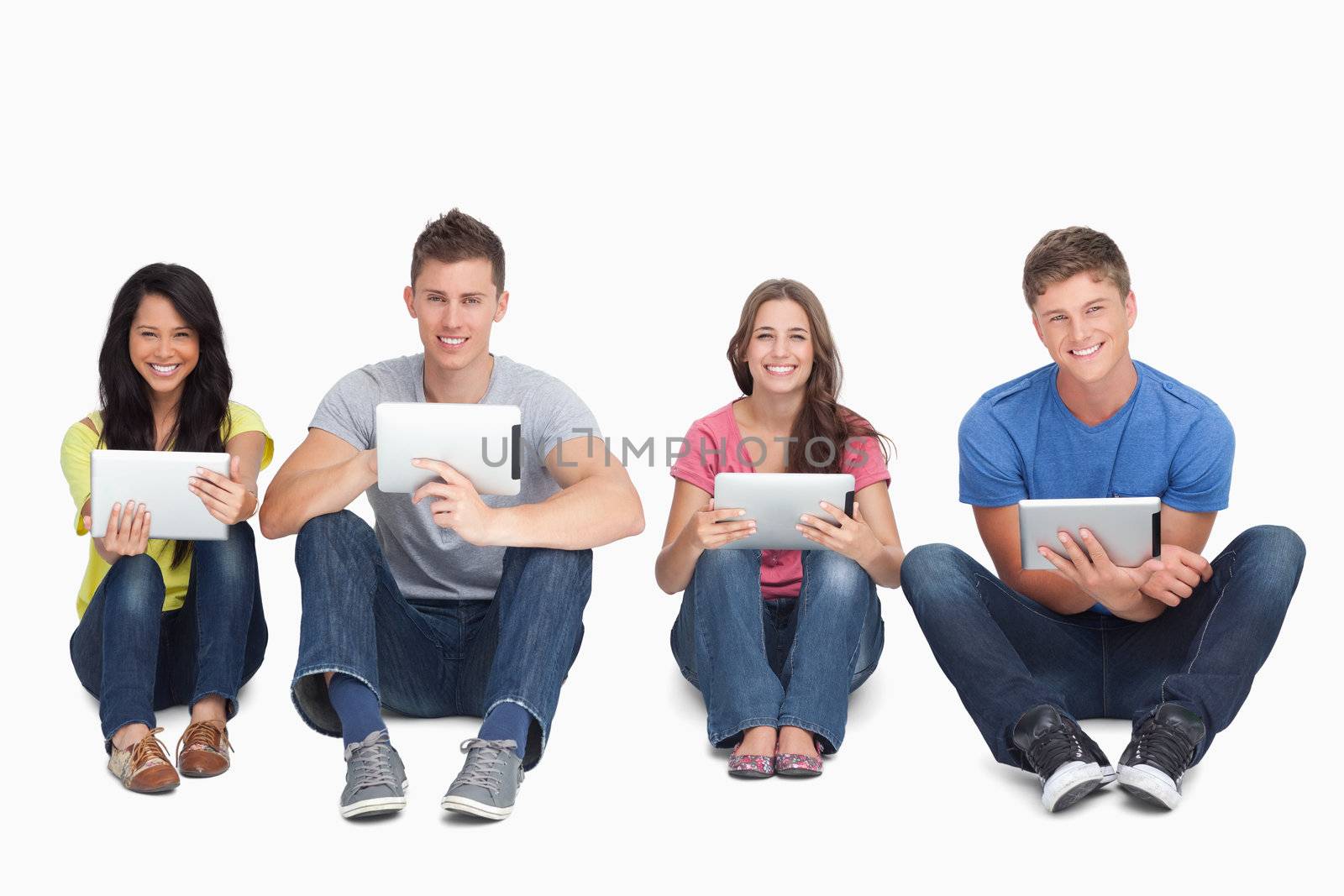 A smiling group of people sitting on the ground beside each other while using tablets and looking at the camera