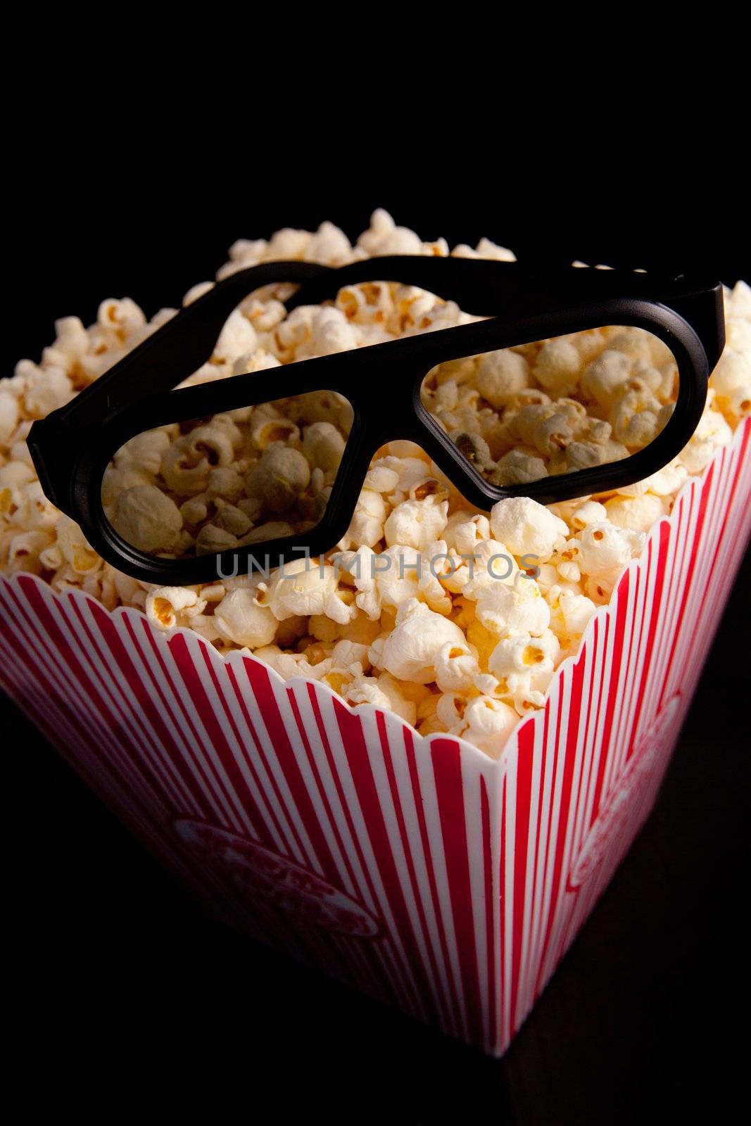 3D glasses lying on the top of a box of popcorn against a black background