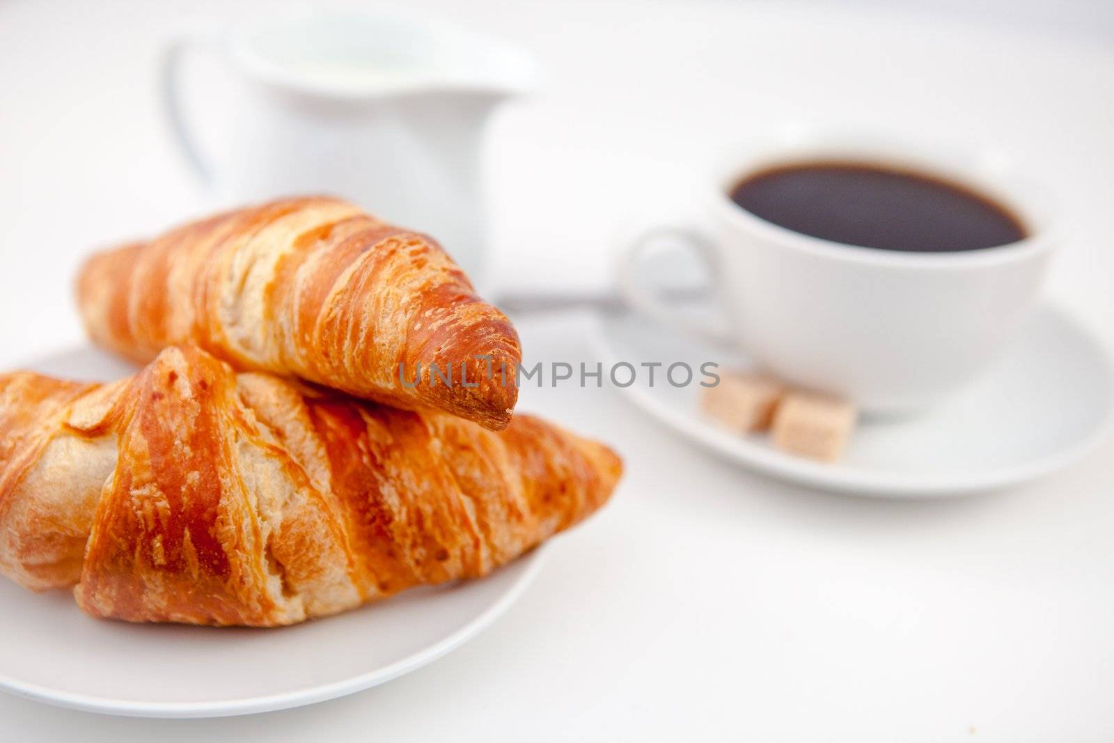 Two croissants and a cup of coffee on white plates with sugar and milk against a white background