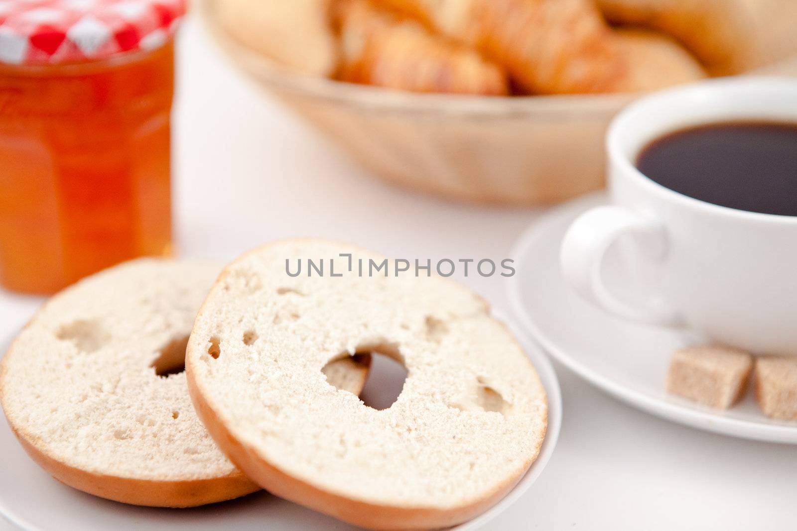 Doughnut cut in half and a cup of coffee on white plates with sugar and milk and a pot of jam against a white background