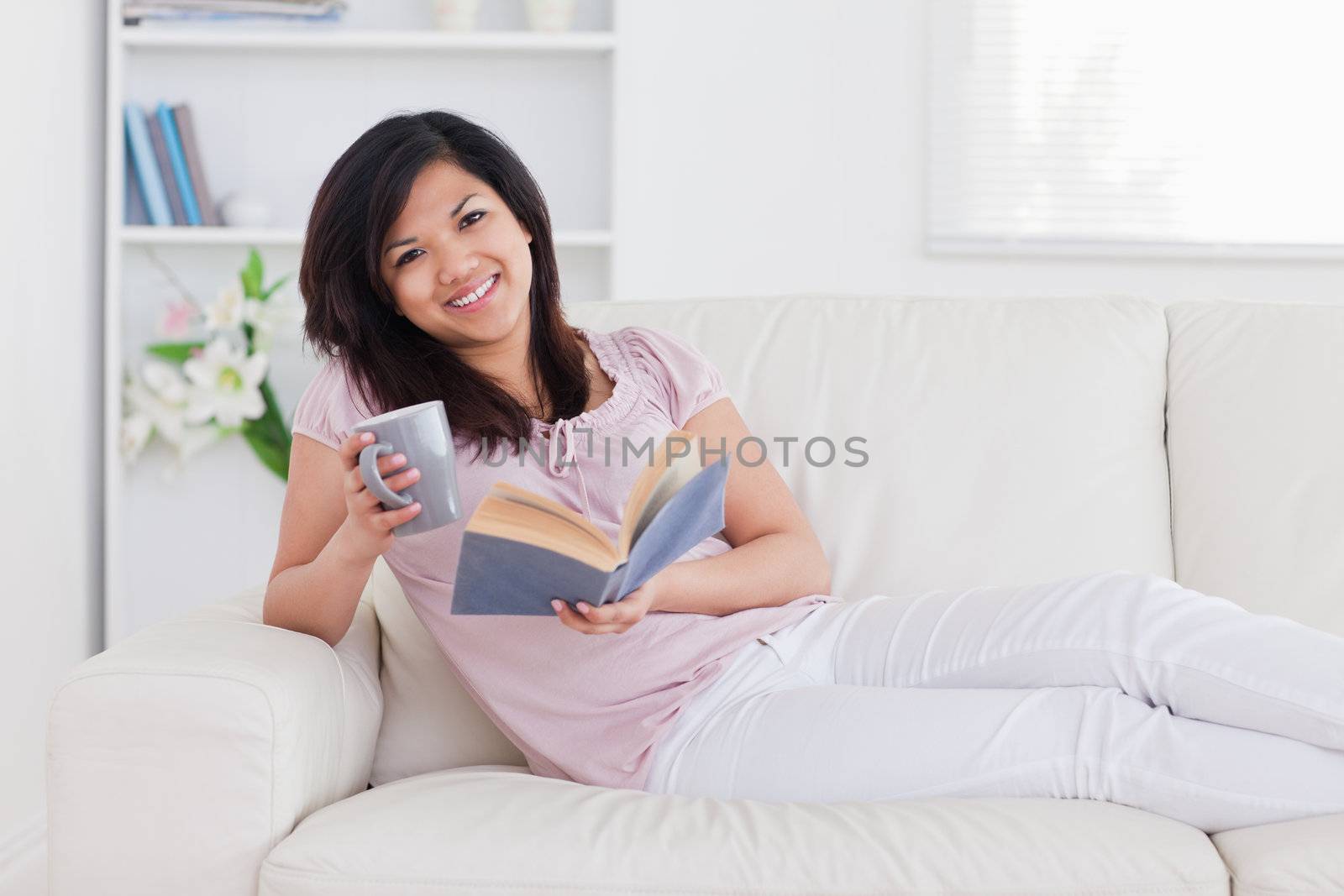 Smiling woman holding a book and a mug while lying on a couch in a living room