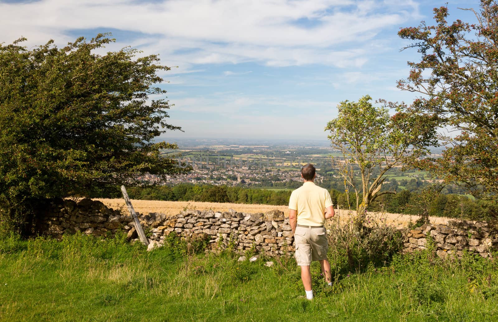 Senior man looks at views over Winchcombe from the barrow at Belas Nap in English Cotswolds