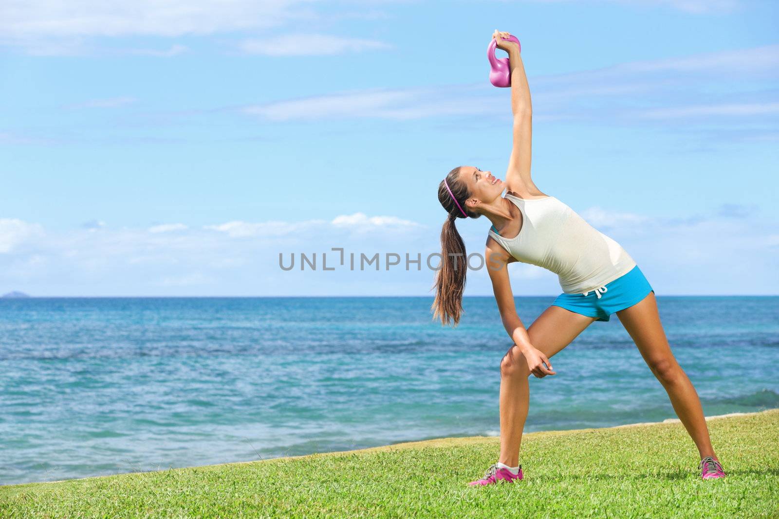 Crossfit exercise woman using kettlebell in fitness strength training workout outside on grass by the ocean. Beautiful young fit instructor and fitness model.