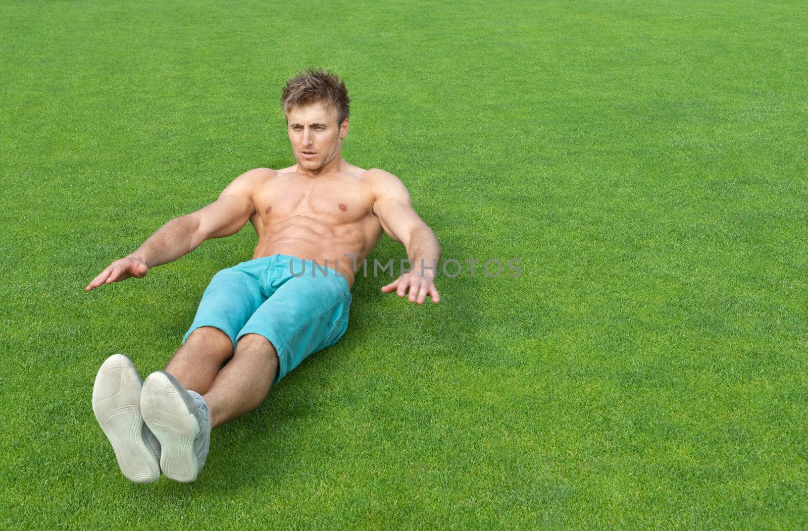 Young man training outdoors and doing sit-ups on green sports field.