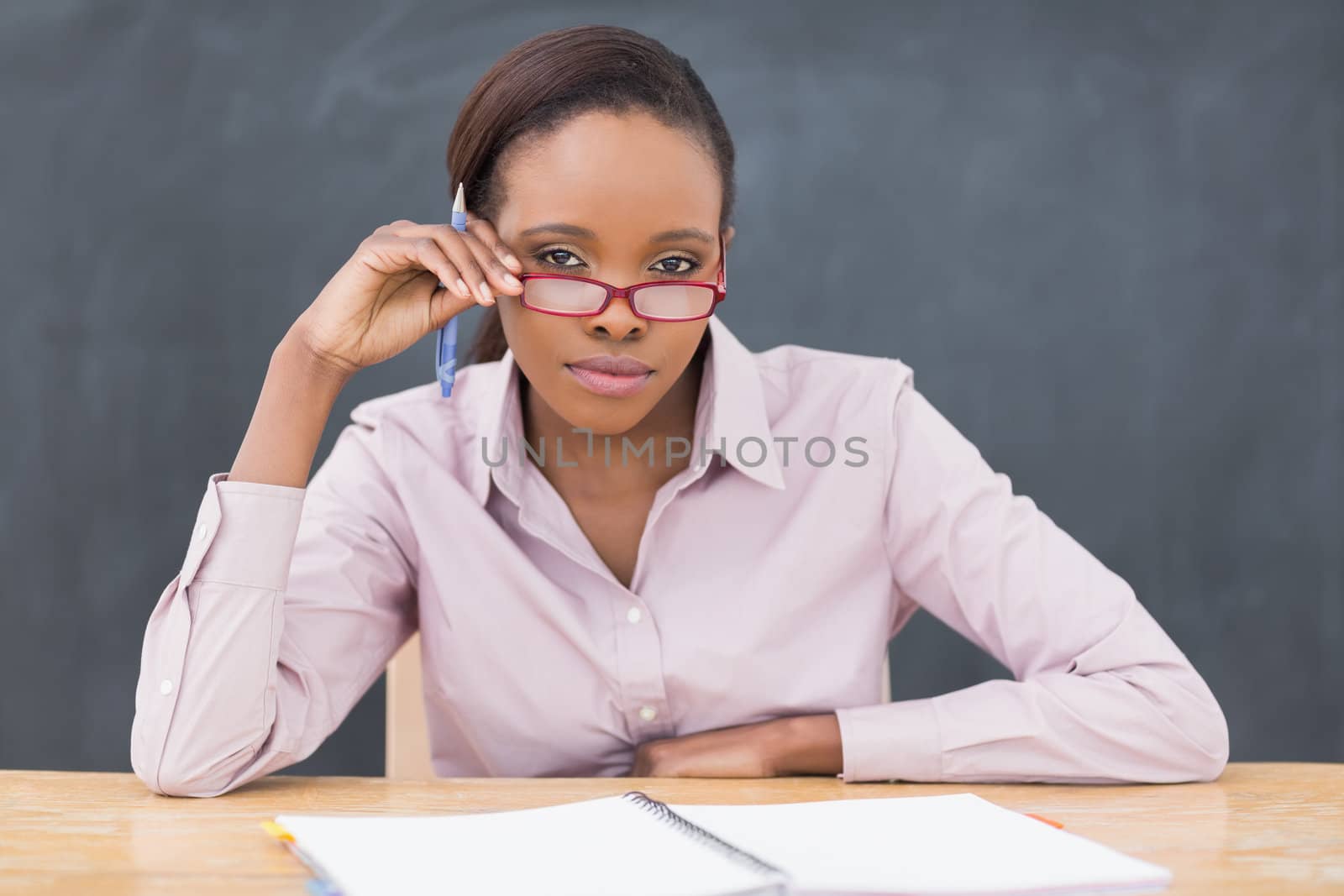 Serious teacher sitting at desk while touching her glasses in a classroom