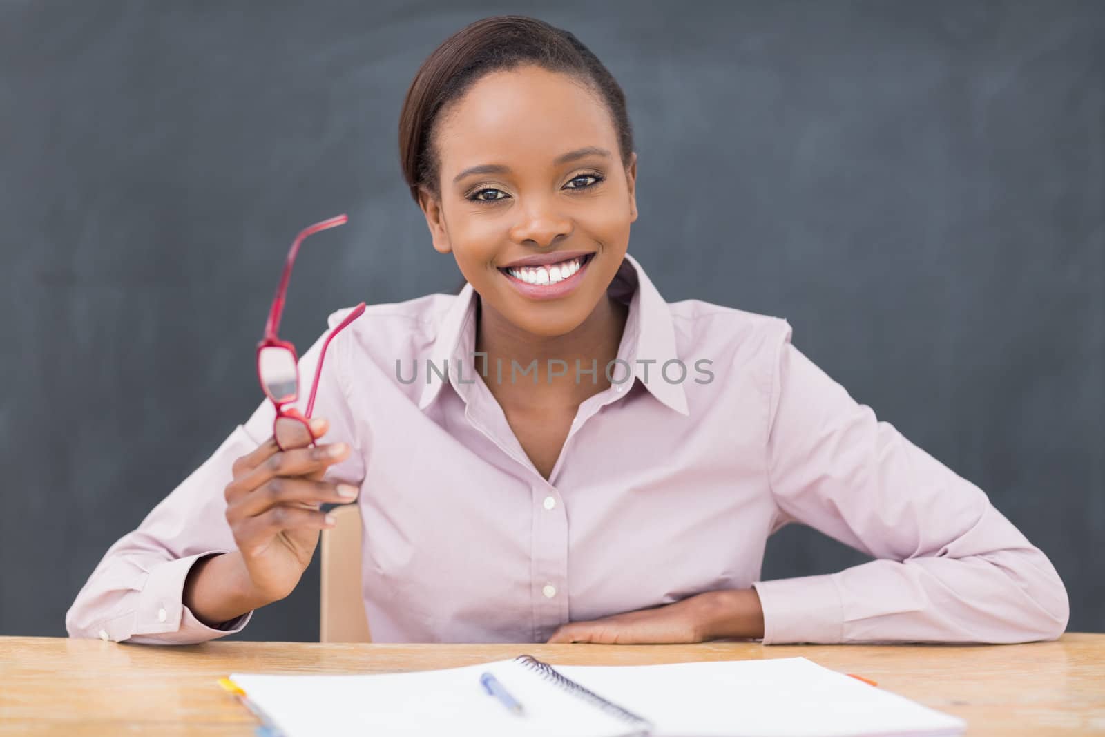 Teacher sitting at desk while holding glasses in a classroom