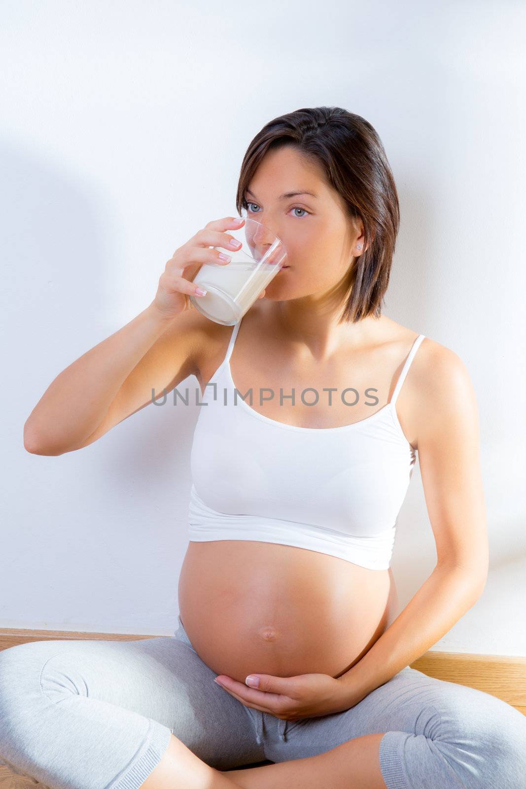 Beautiful pregnant woman drinking milk at home sitting on wooden floor
