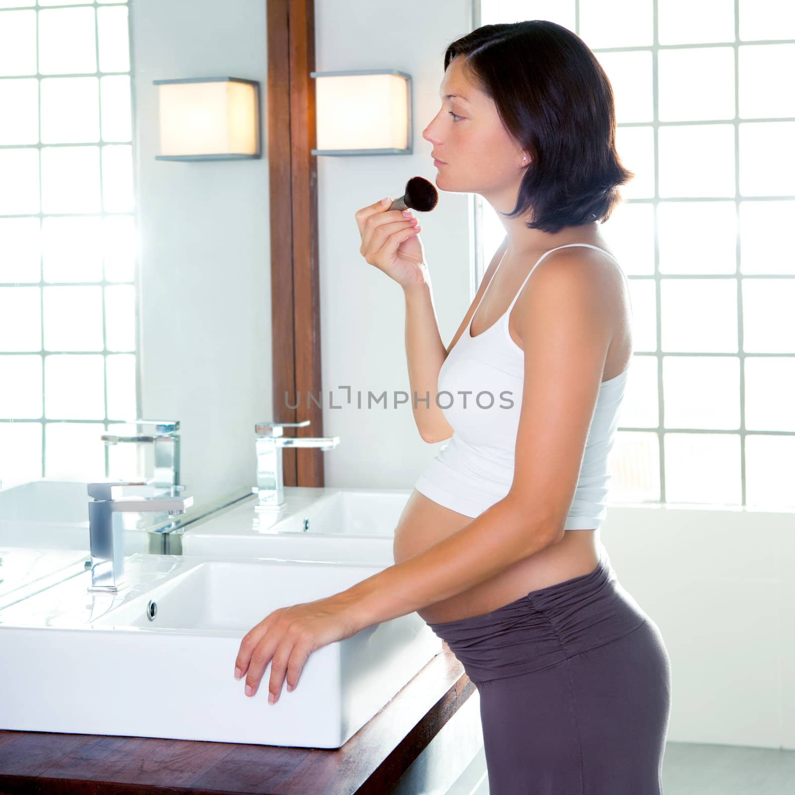 Beautiful pregnant woman on modern bathroom makeup with brush in mirror