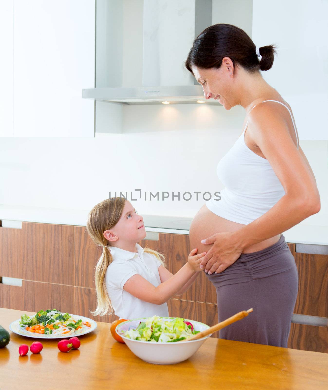 Beautiful pregnant mother with her daughter at kitchen preparing salad