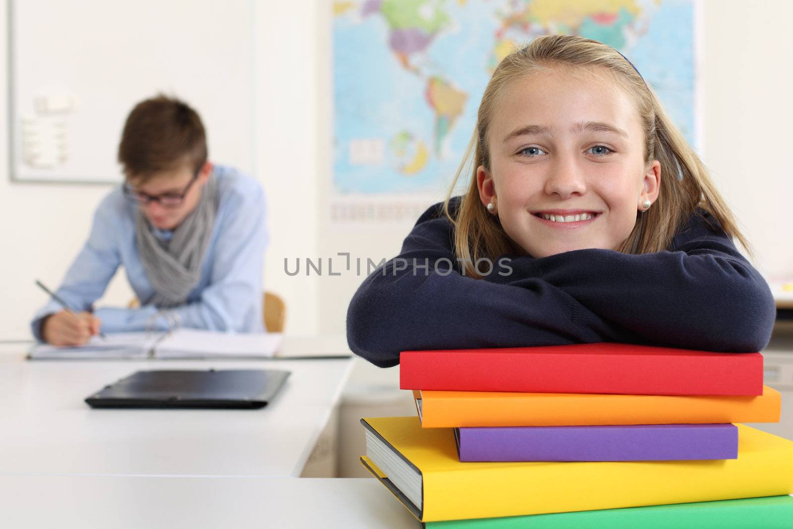 Photo of a pretty young girl smiling in her classroom with a male in the background writing and reading.