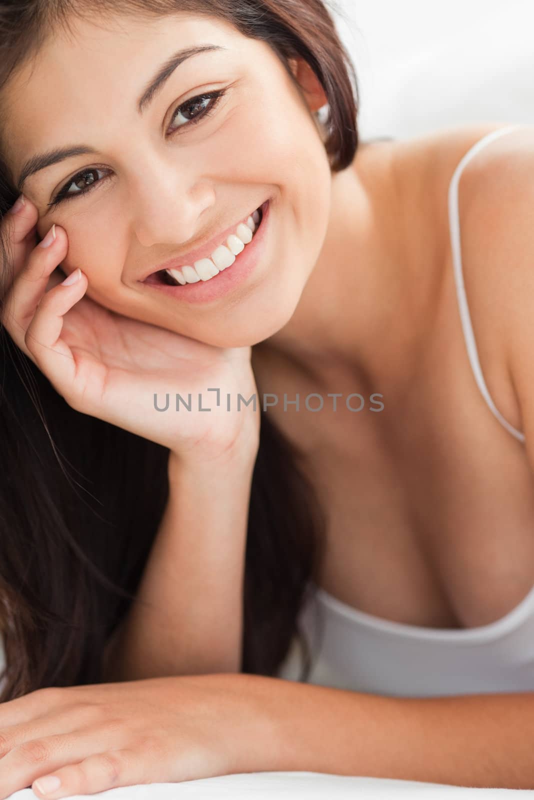 A close up shot of a smiling woman who is looking forward with a hand resting on her cheek and her other hand upon a pillow.