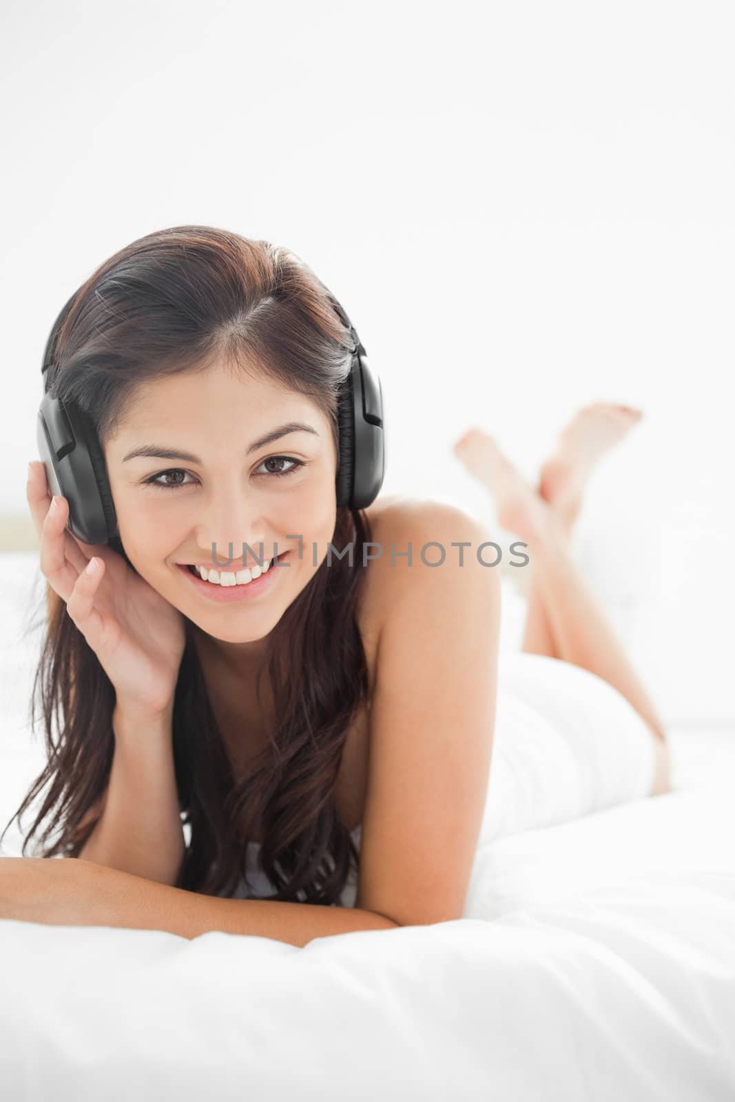 A close up picture of a  smiling woman with her headphones on, with her hand on them, while her legs are crossed.