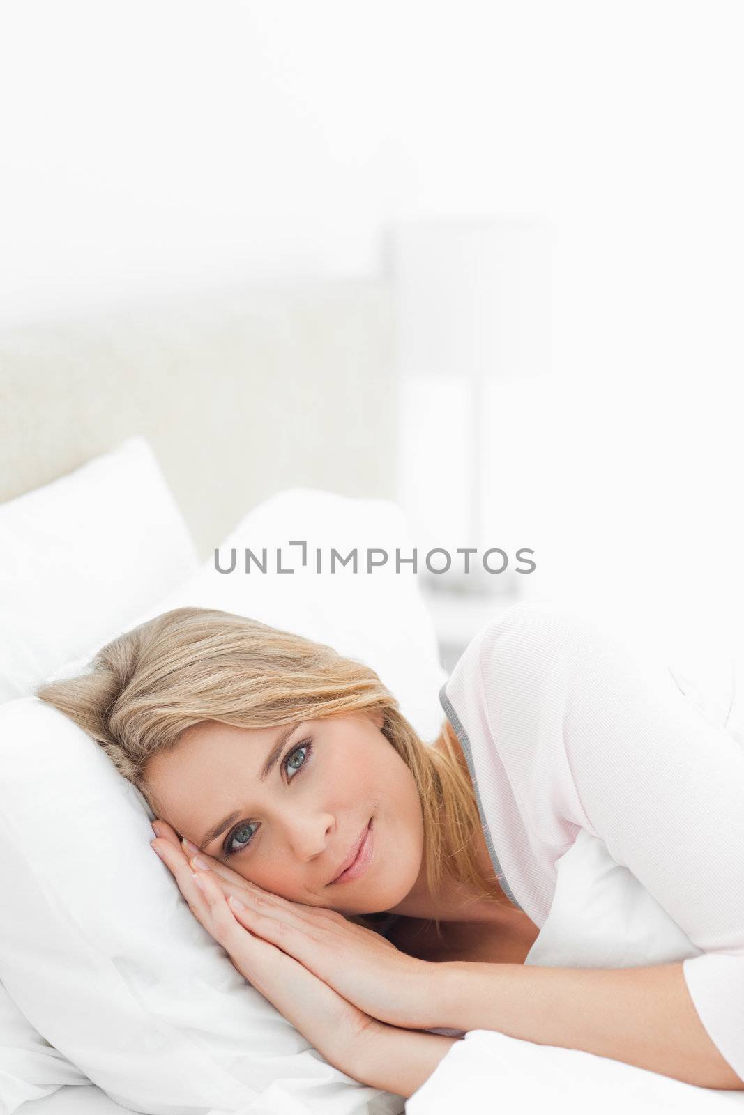 A woman resting in the bed, with her eyes open and her hands and head both resting on the pillow.