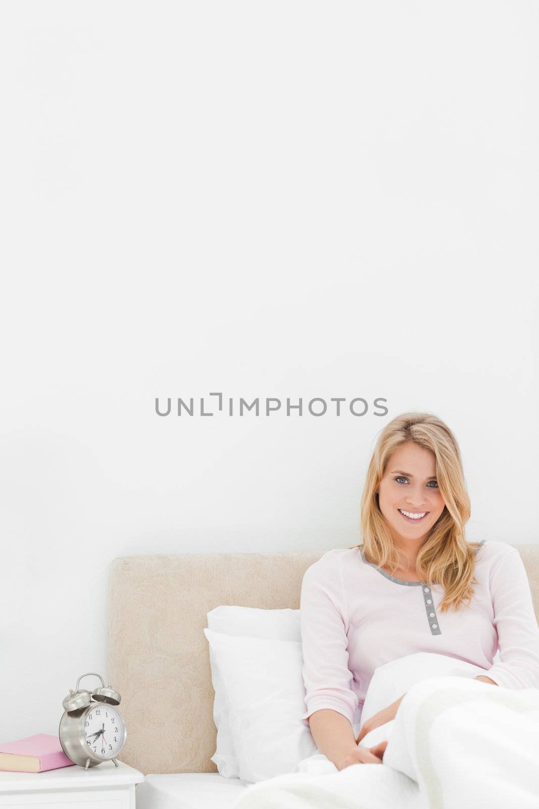 A Vertical shot of a smiling woman sitting in bed against the headboard with the alarm clock on a desk beside her.