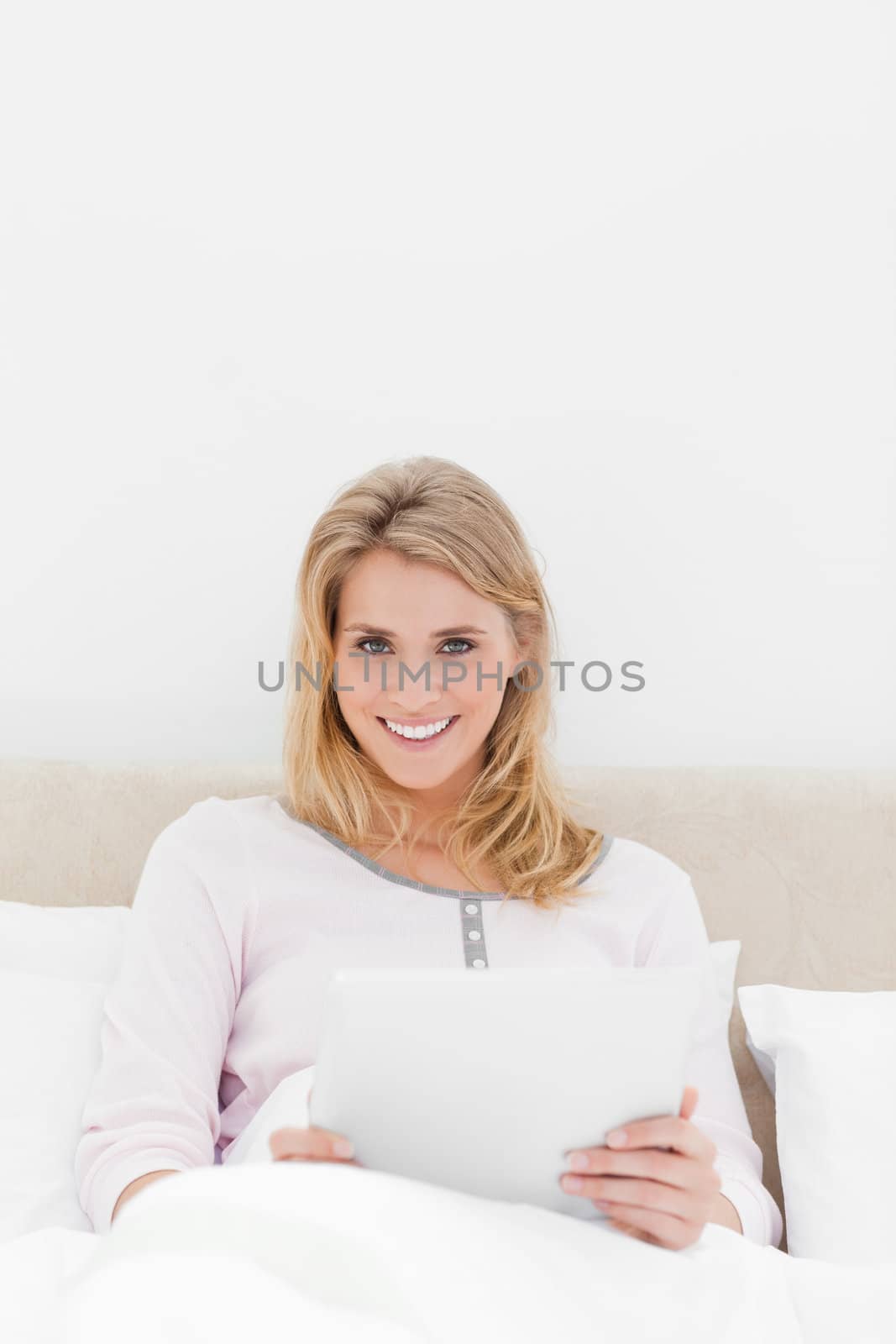 A centered shot of a woman with a tablet pc, looking forward and smiling while in bed.