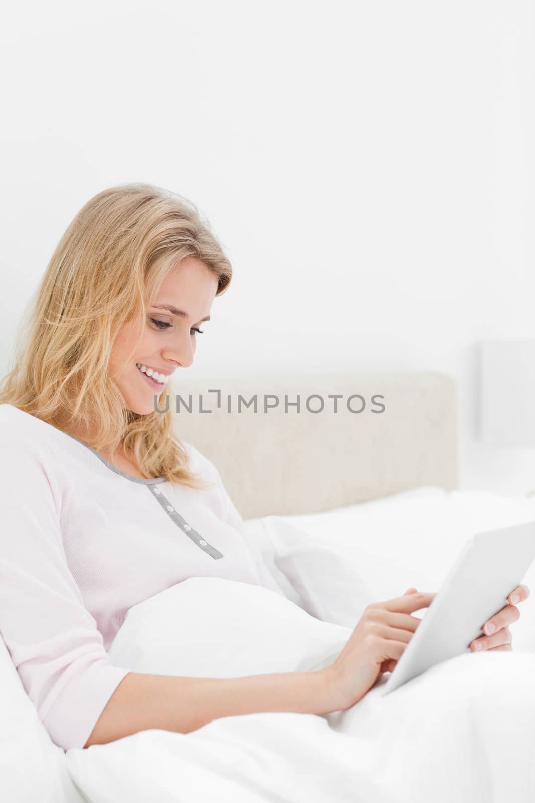 A side view shot of a woman sitting in bed with a tablet pc that she is watching while smiling.