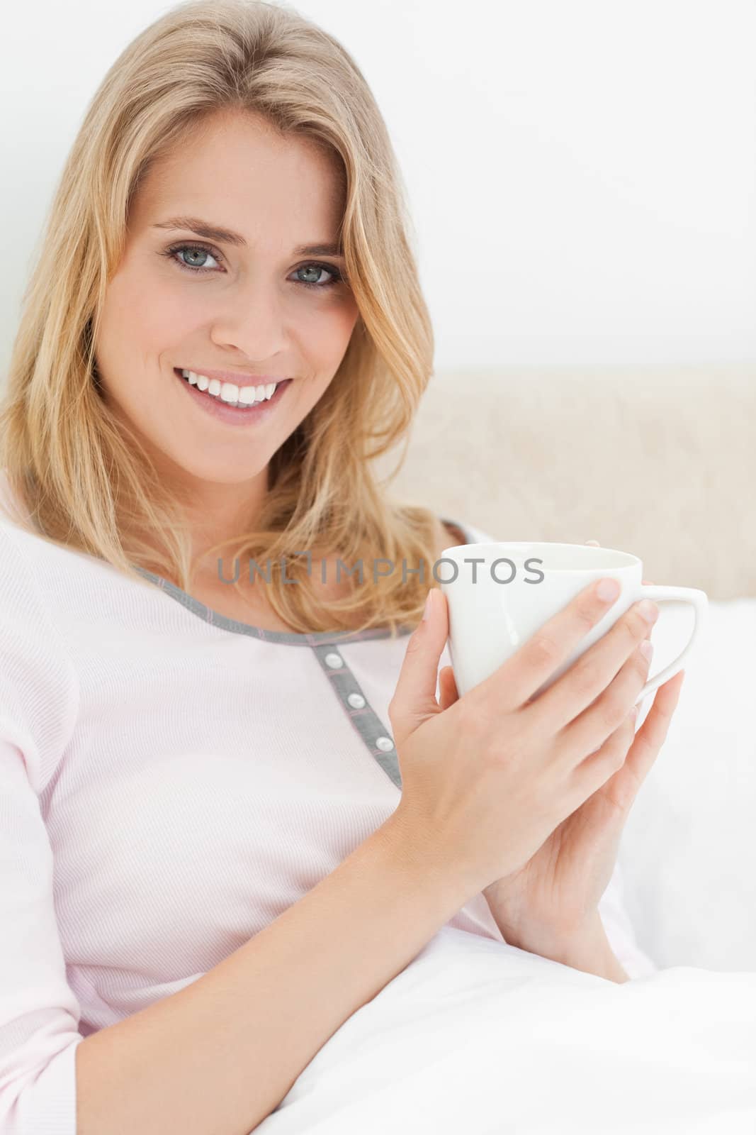 A close up shot of a woman holding a cup in her hands near to her head as she looks forward with a smile.