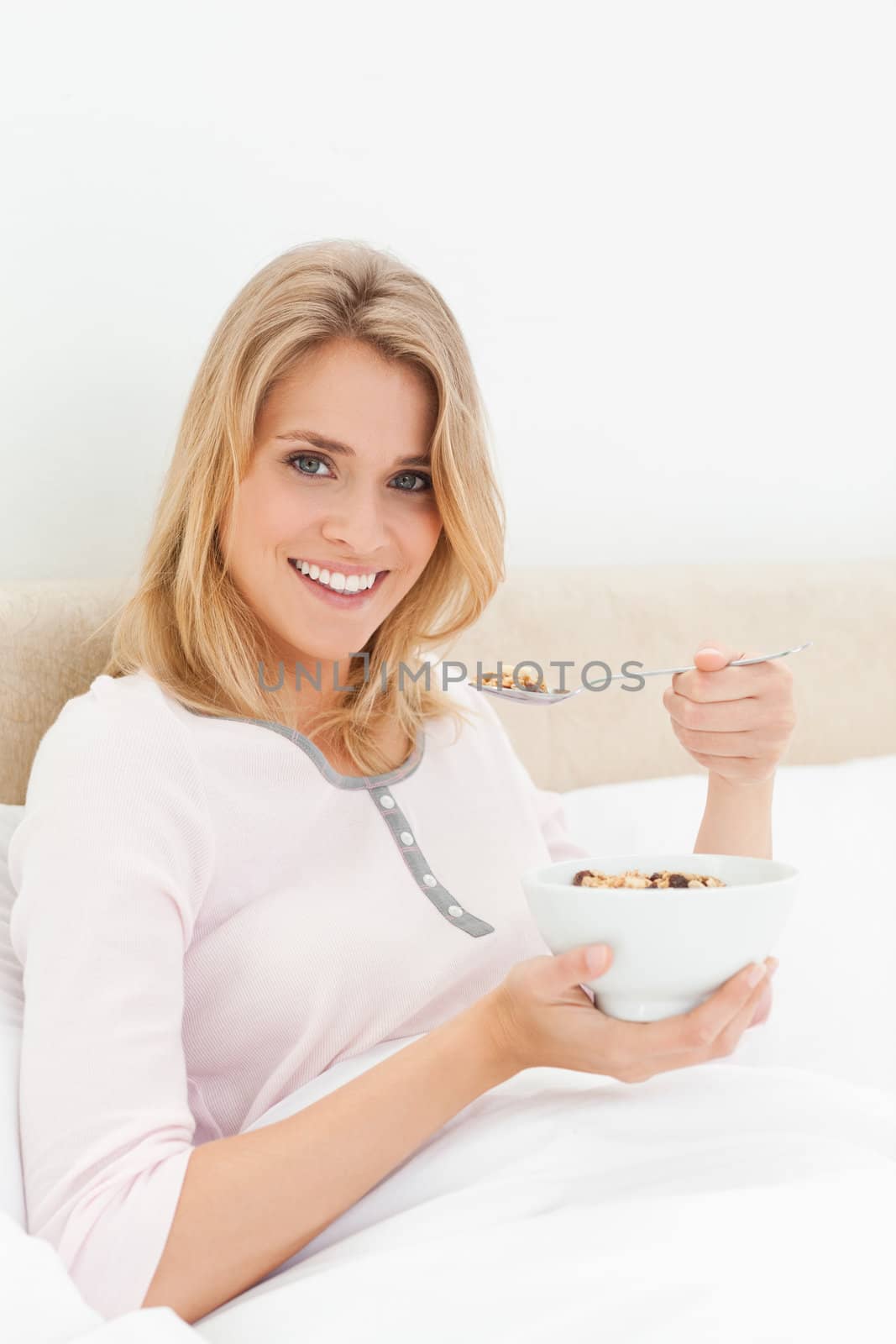 A woman sitting in bed with a bowl of cereal in hand and a spoon of cereal near her mouth, while looking ahead and smiling.