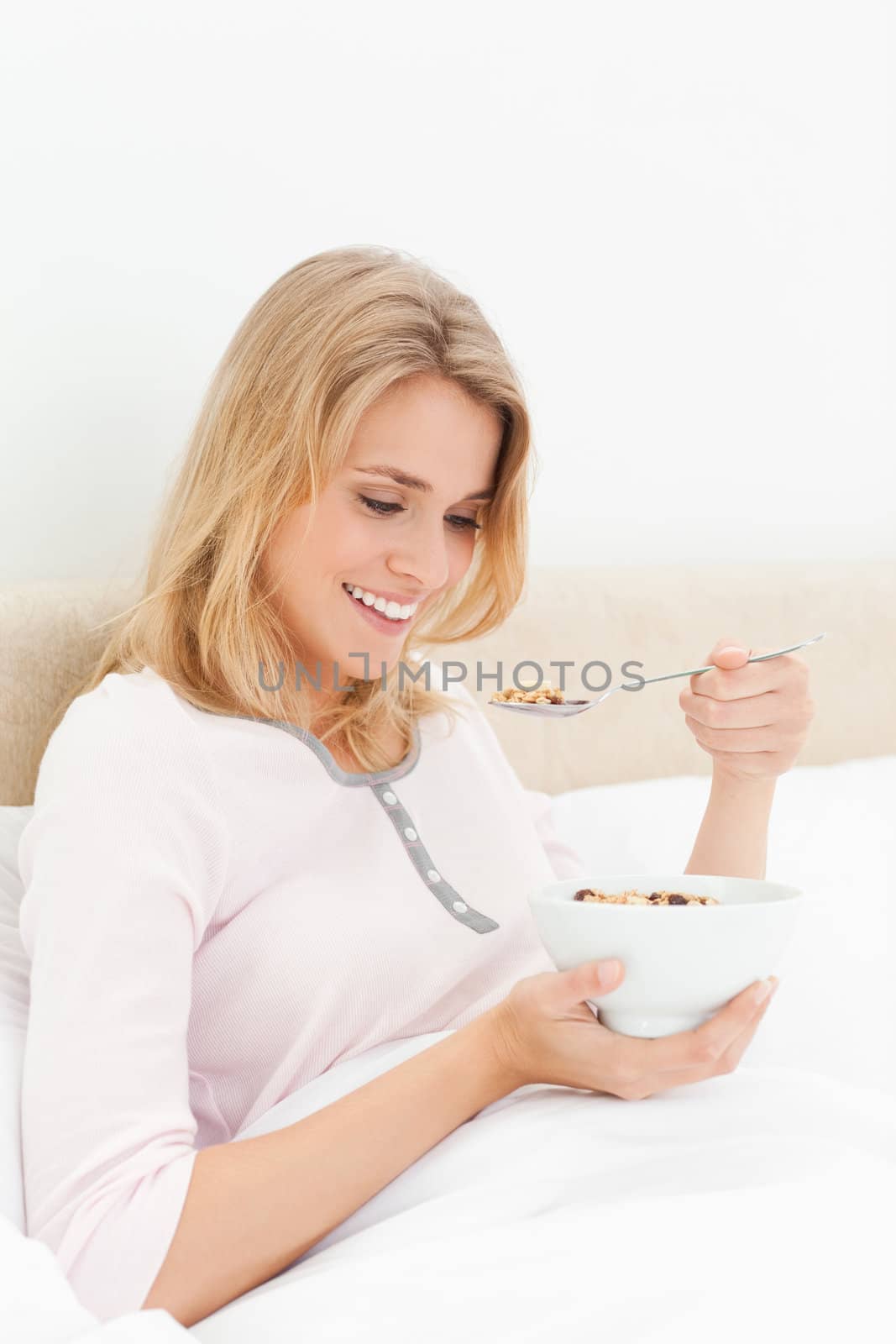 A woman sitting in bed with a bowl of cereal in her and and a spoon of cereal near her mouth as she looks at it and smiles.