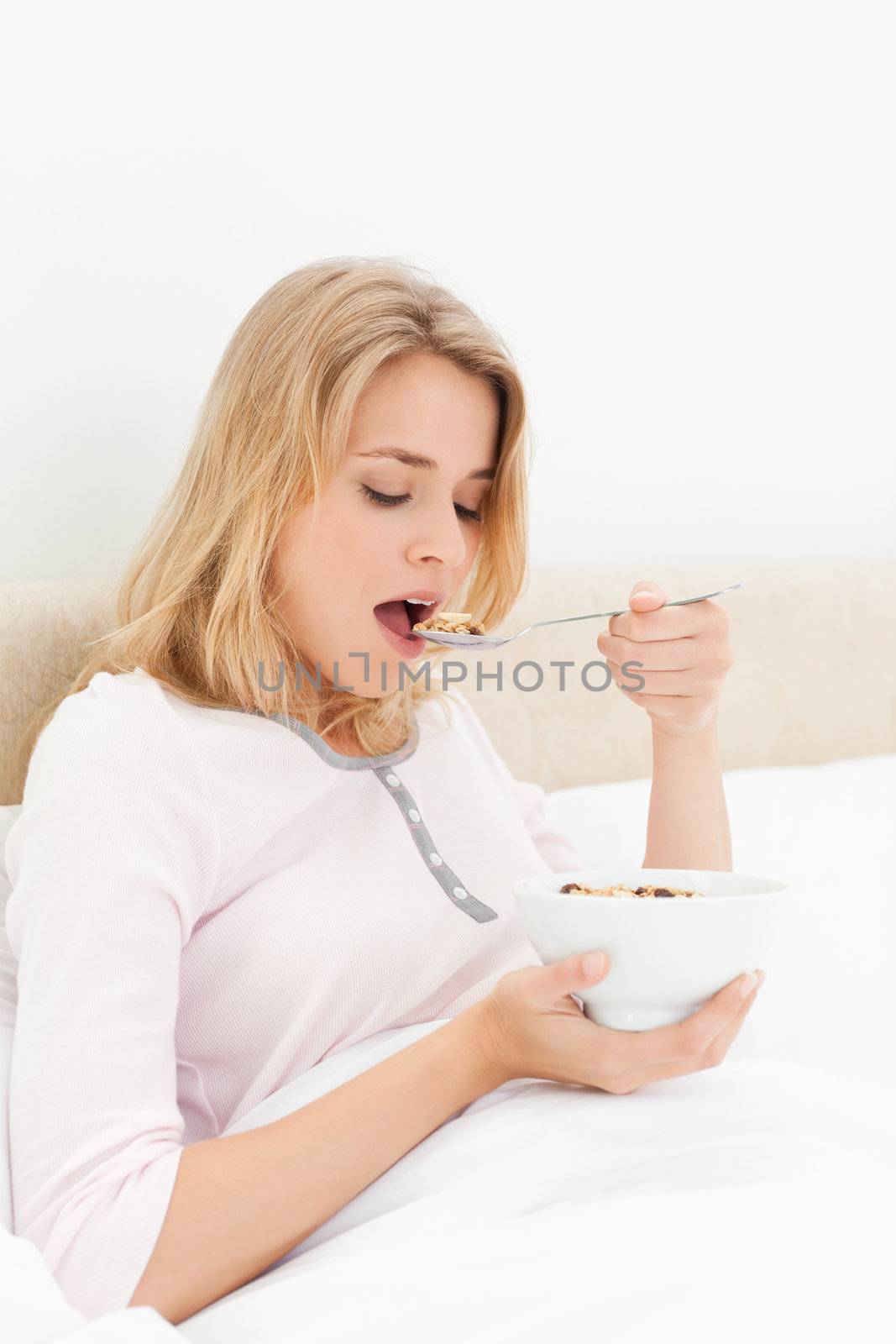 A woman sitting in bed, with a bowl in hand, and about to eat a spoon of cereal.