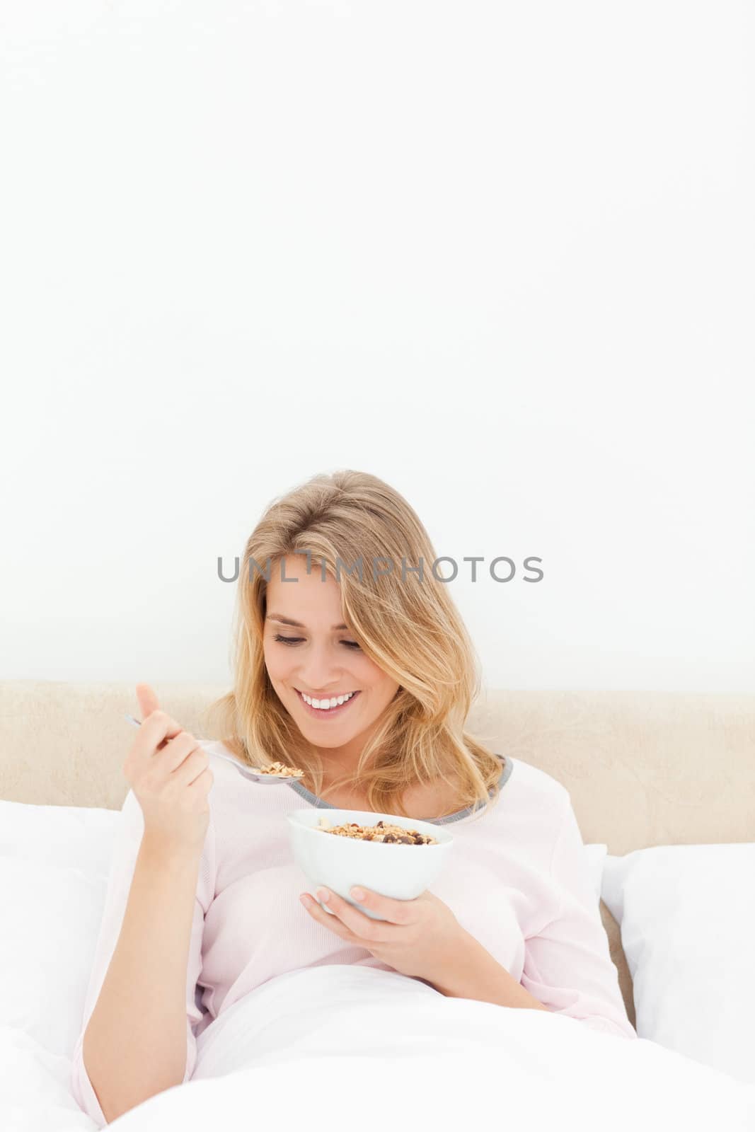 A vertical shot of a woman in bed with a bowl of cereal, as she smiles and looks at the spoon of cereal.