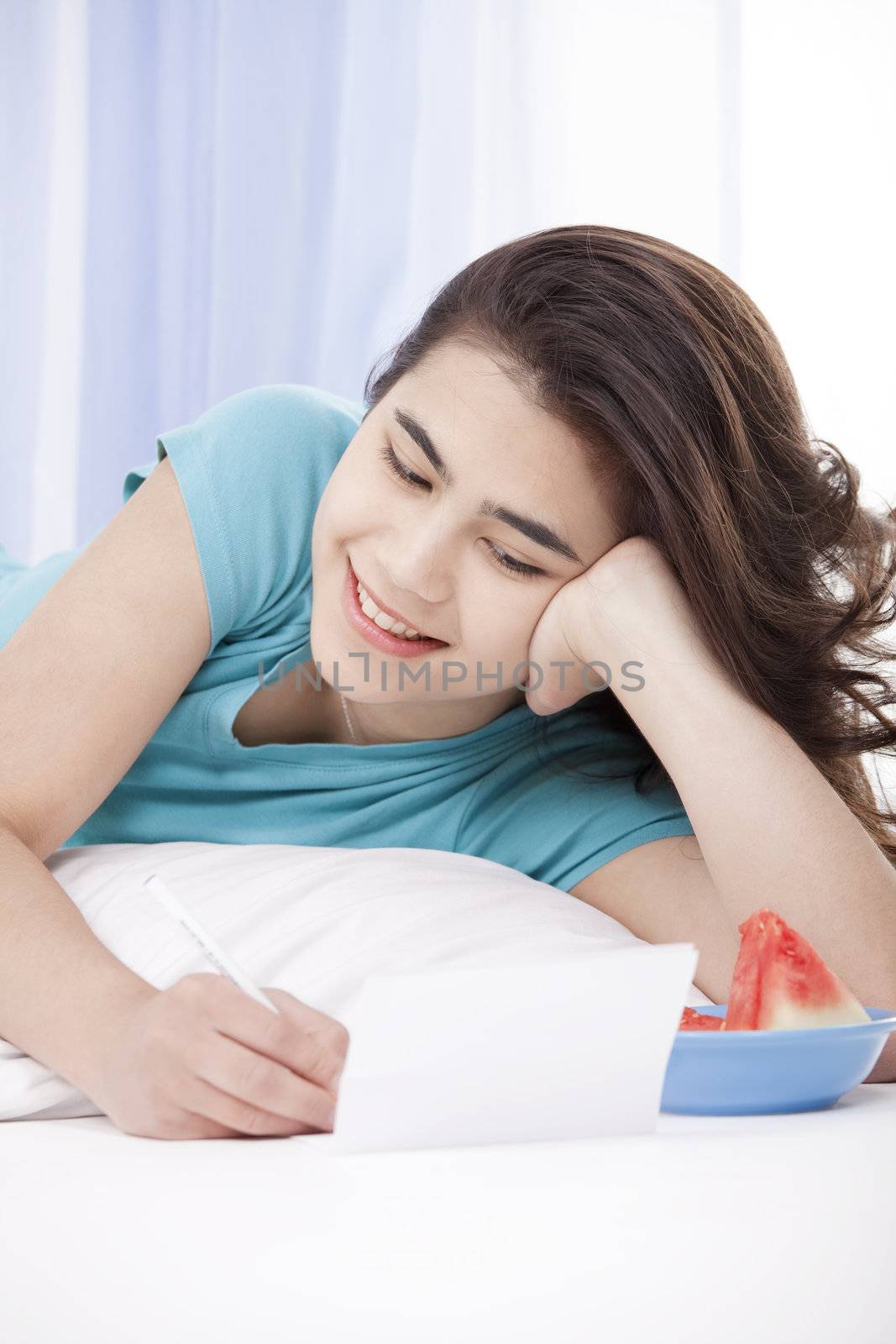 Relaxed and happy biracial teen girl or young woman lying on floor pillow writing a letter or note.
