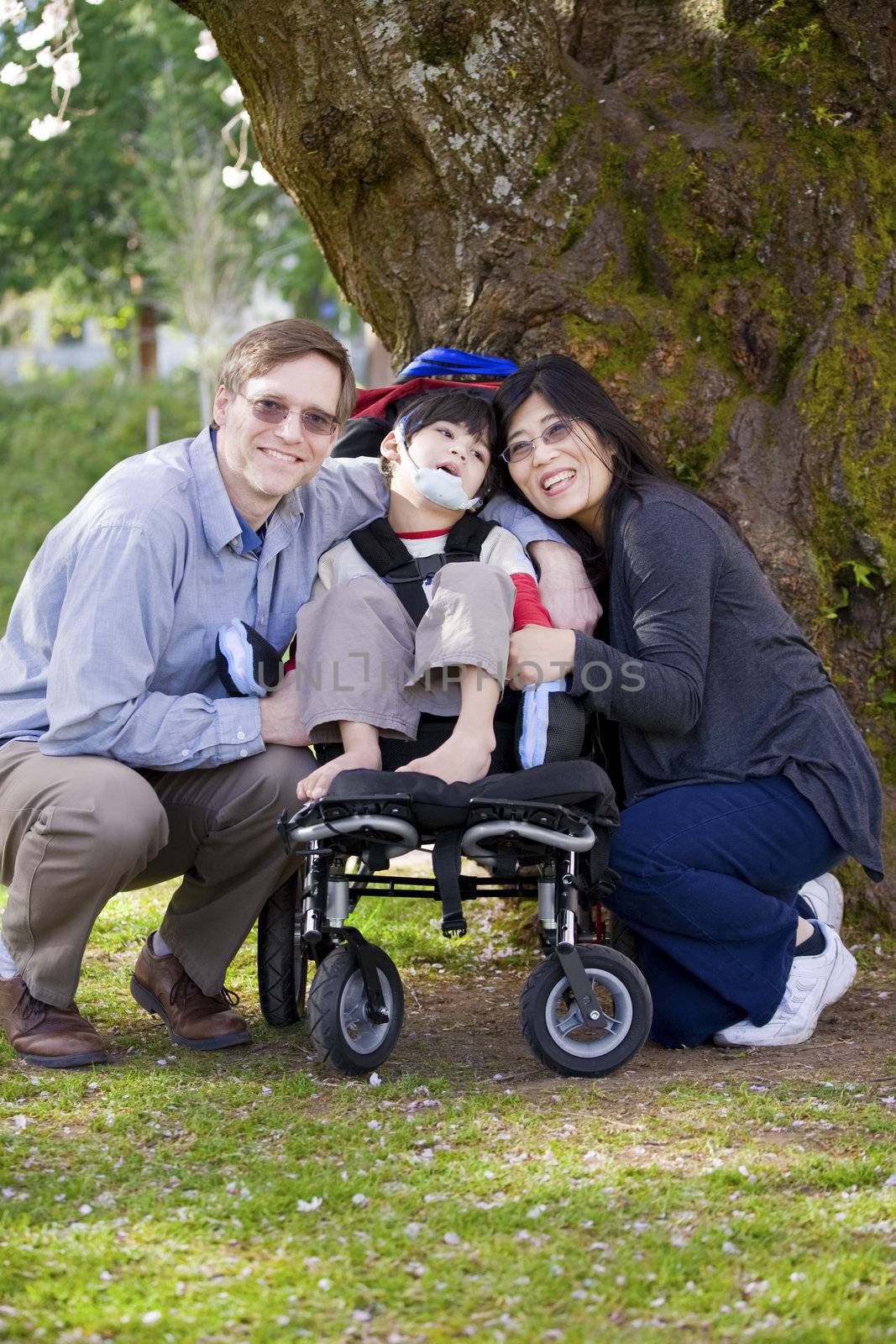 Happy disabled child in wheelchair surrounded by parents, outdoors. Cerebral palsy.