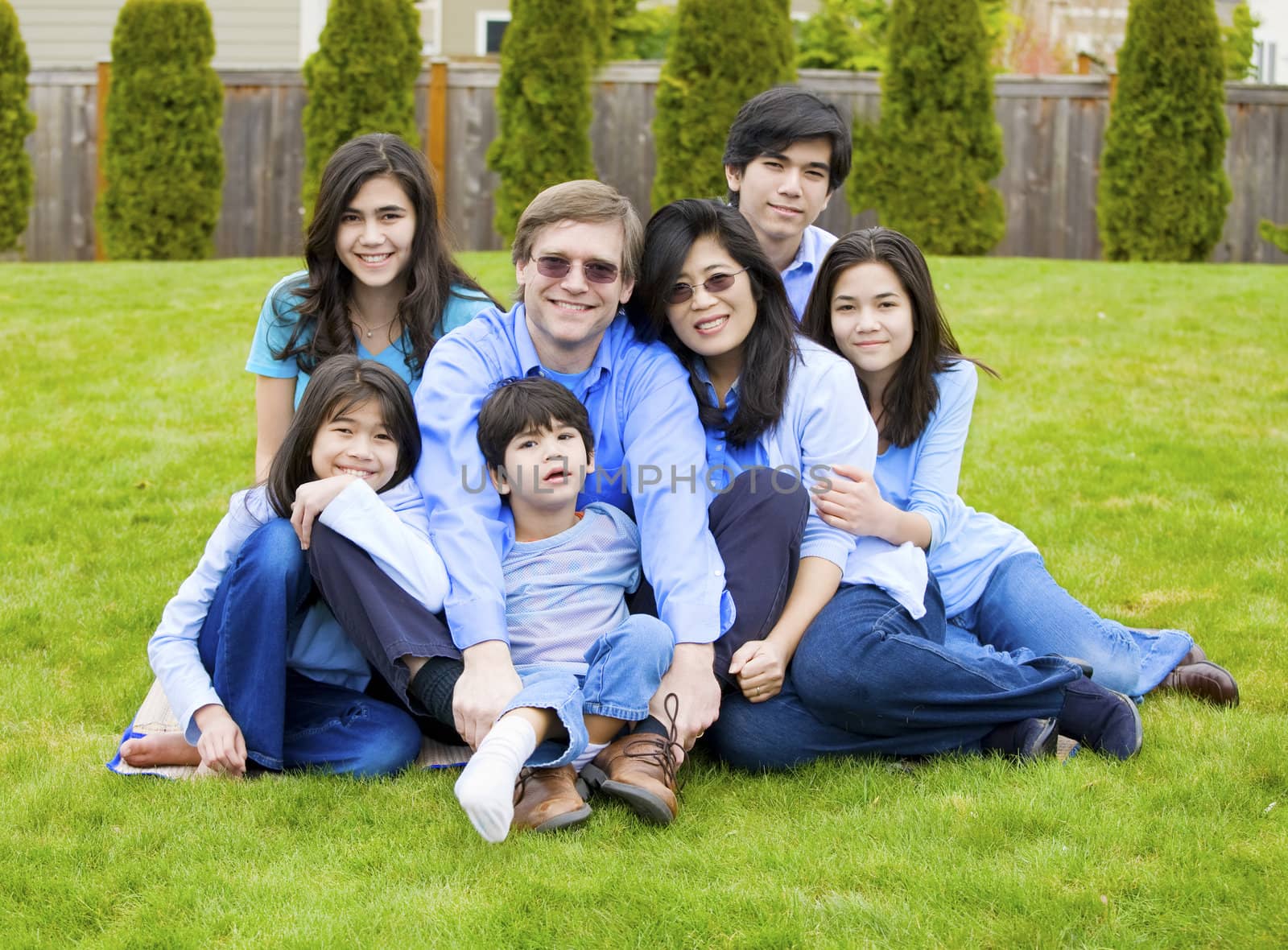 Large multiracial family of seven sitting together on lawn, dressed in blue colors. Five year old boy in front is disabled with cerebral palsy.