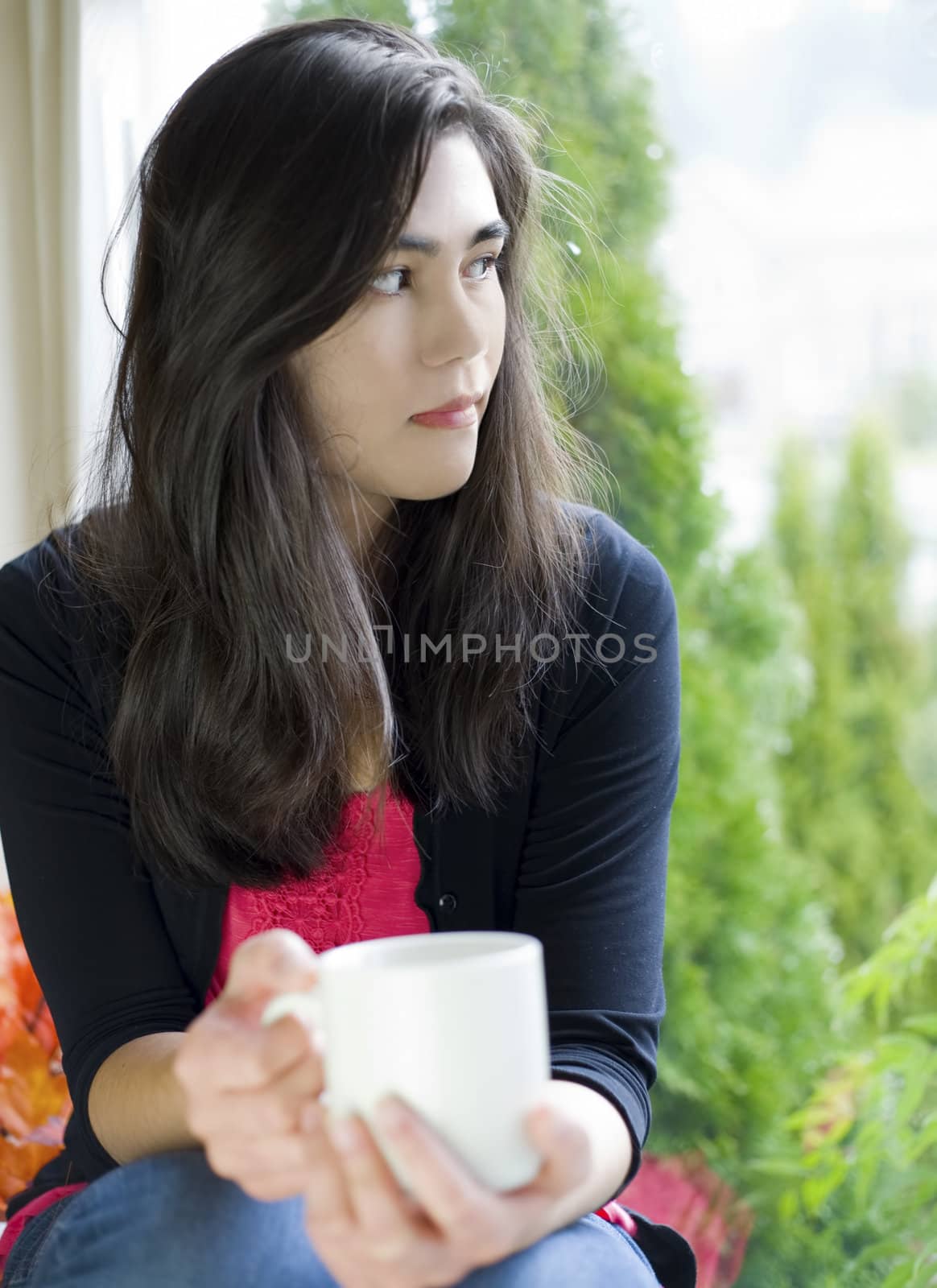 Beautiful biracial teenage girl or young woman holding coffee cup by window, sad or thoughtful expression