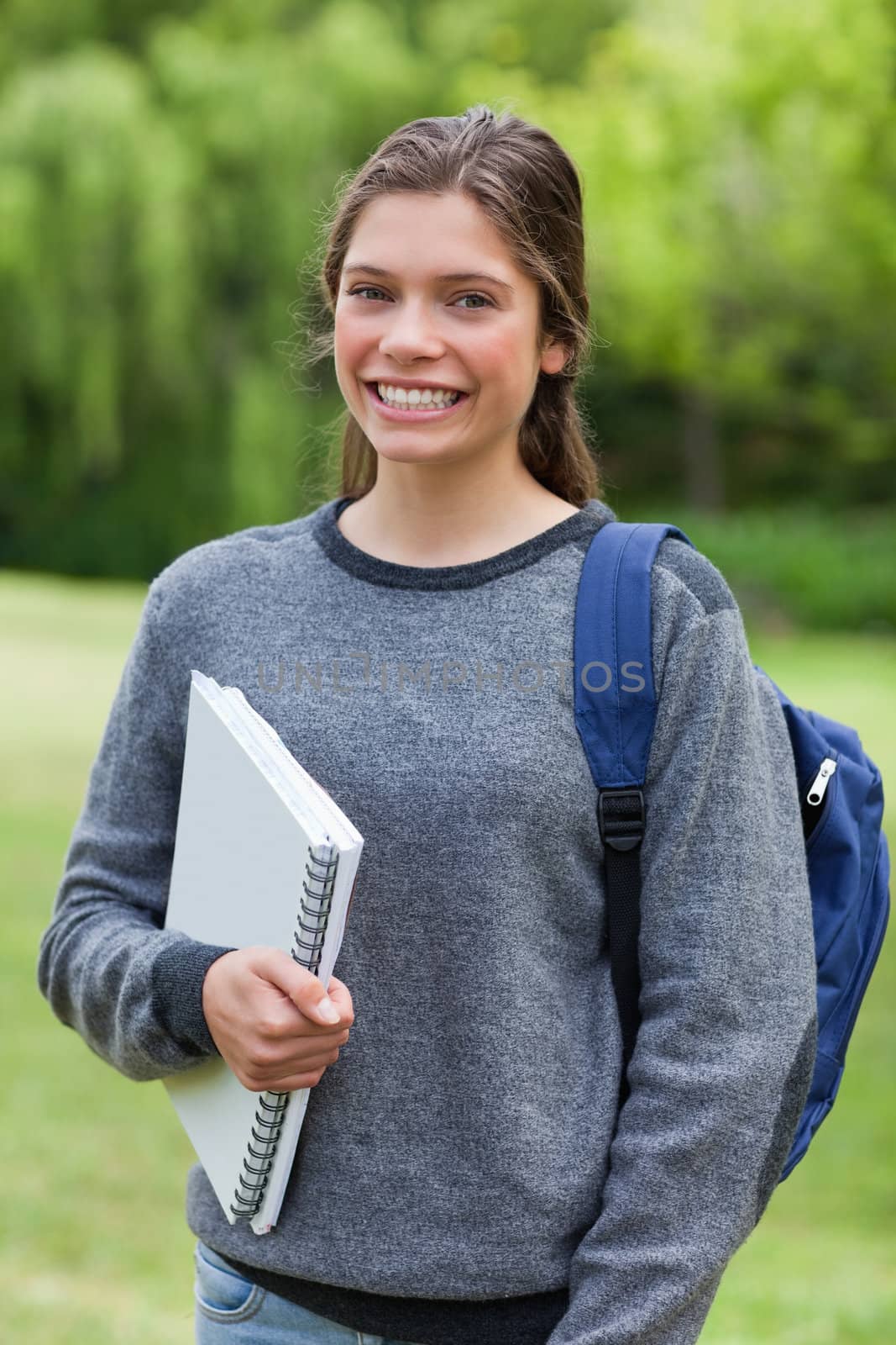 Teenager beaming while holding a notebook and standing upright in a park