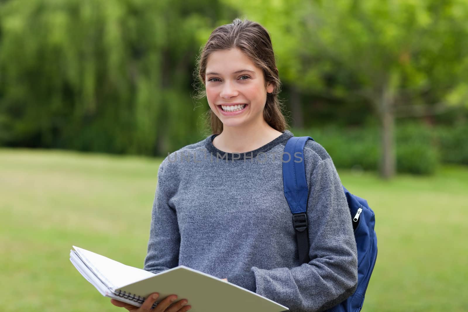 Young girl standing in a park while holding her notebook and beaming