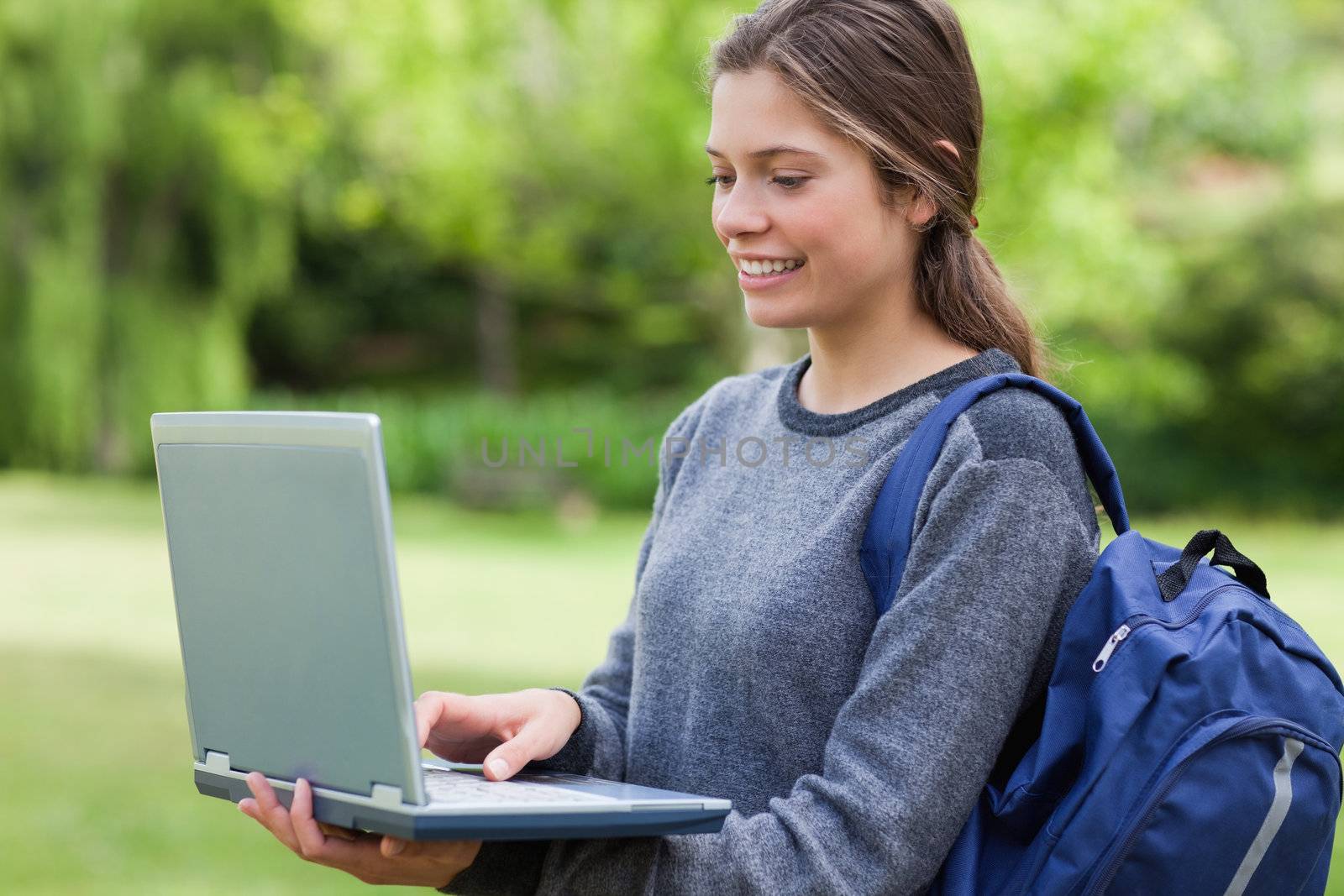 Young smiling woman holding her laptop while standing in a park with hair tied