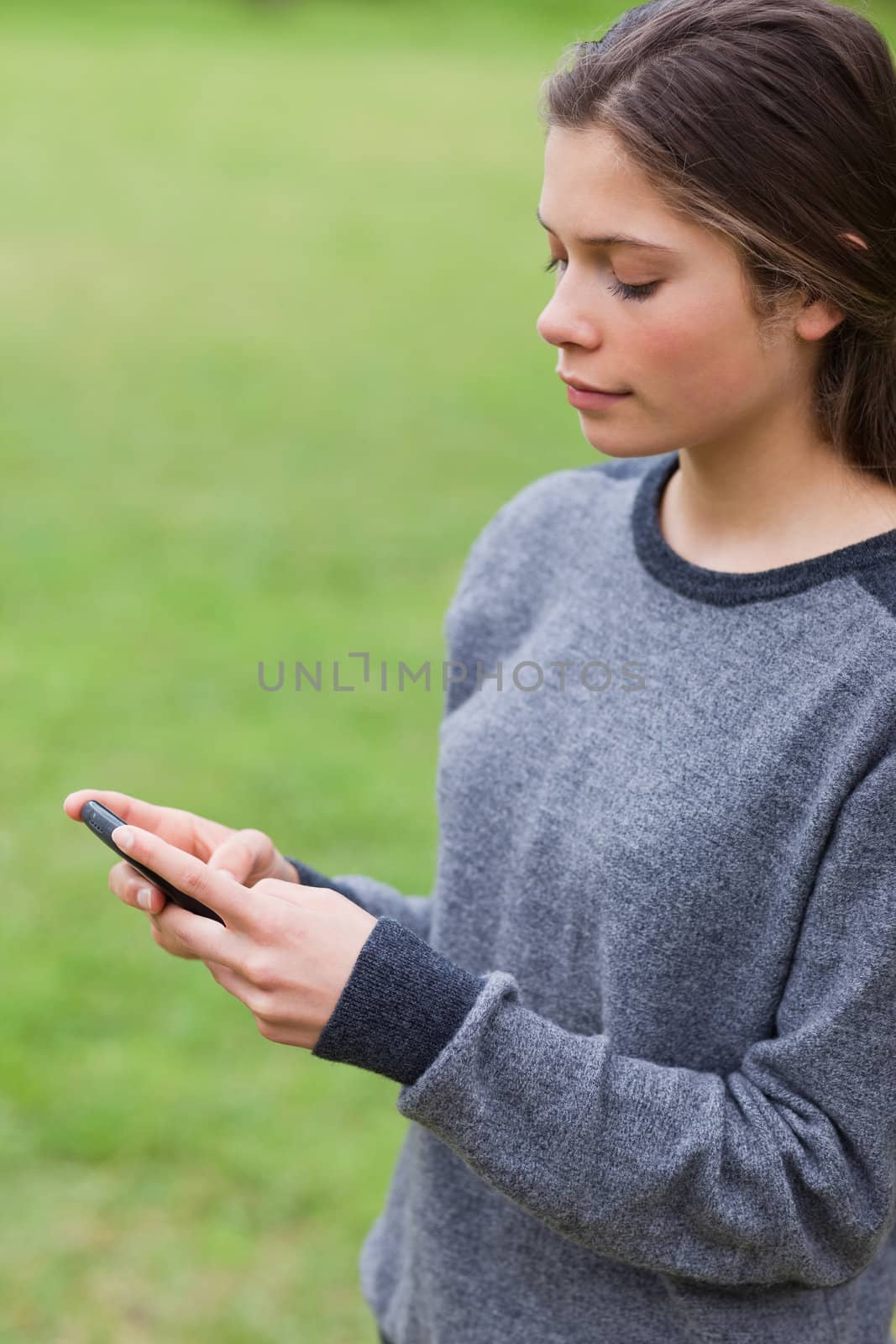 Young serious girl sending a text with her mobile phone while standing in a park