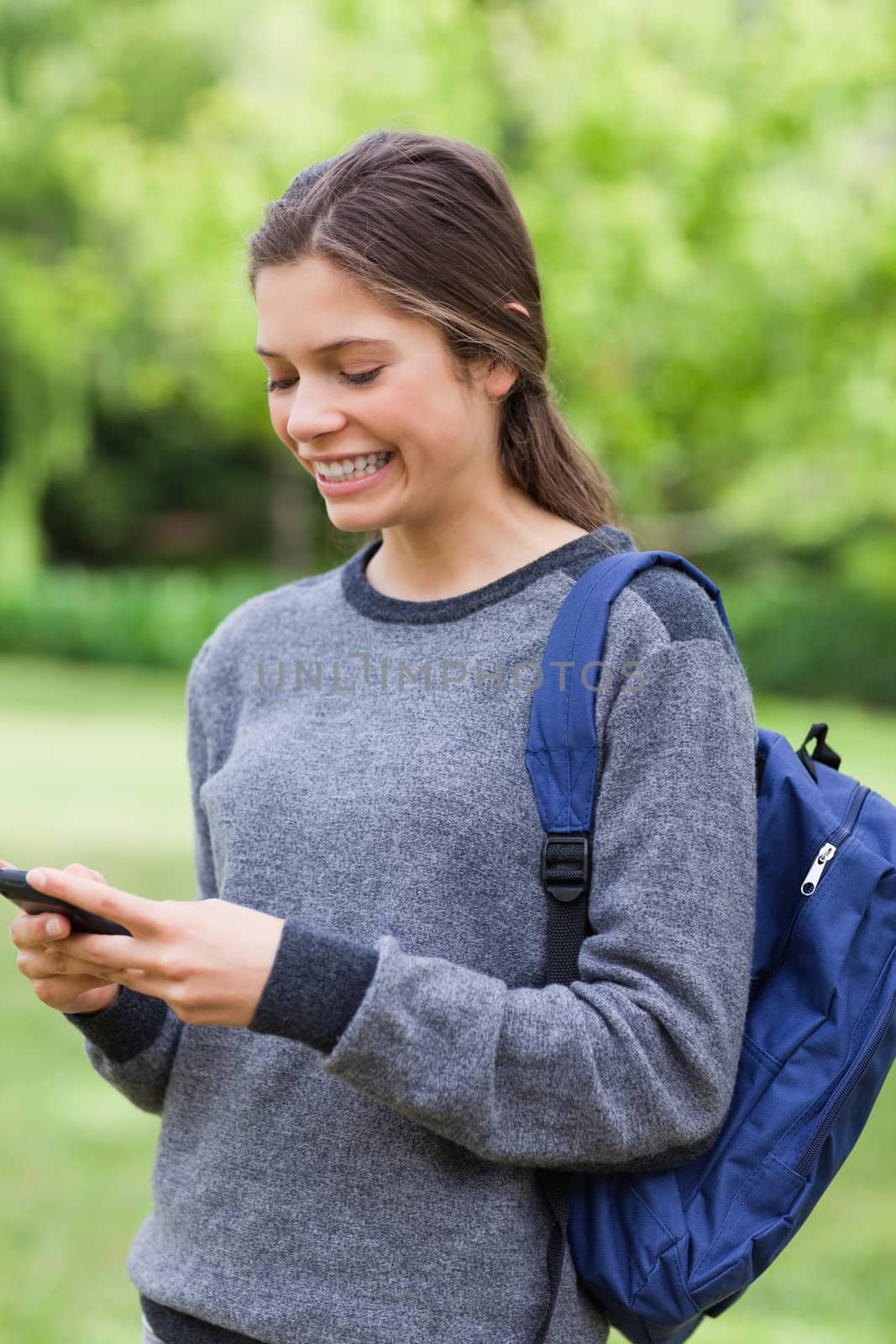 Smiling young girl sending a text with her mobile phone while standing in a park