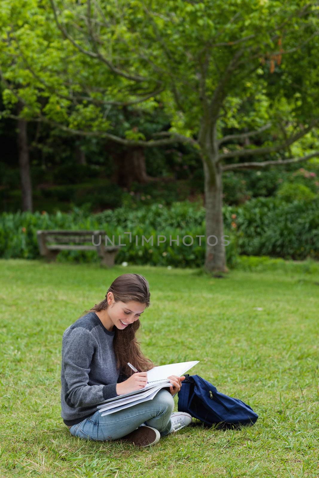 Smiling young adult seriously writing on her notebook while sitting down in a park