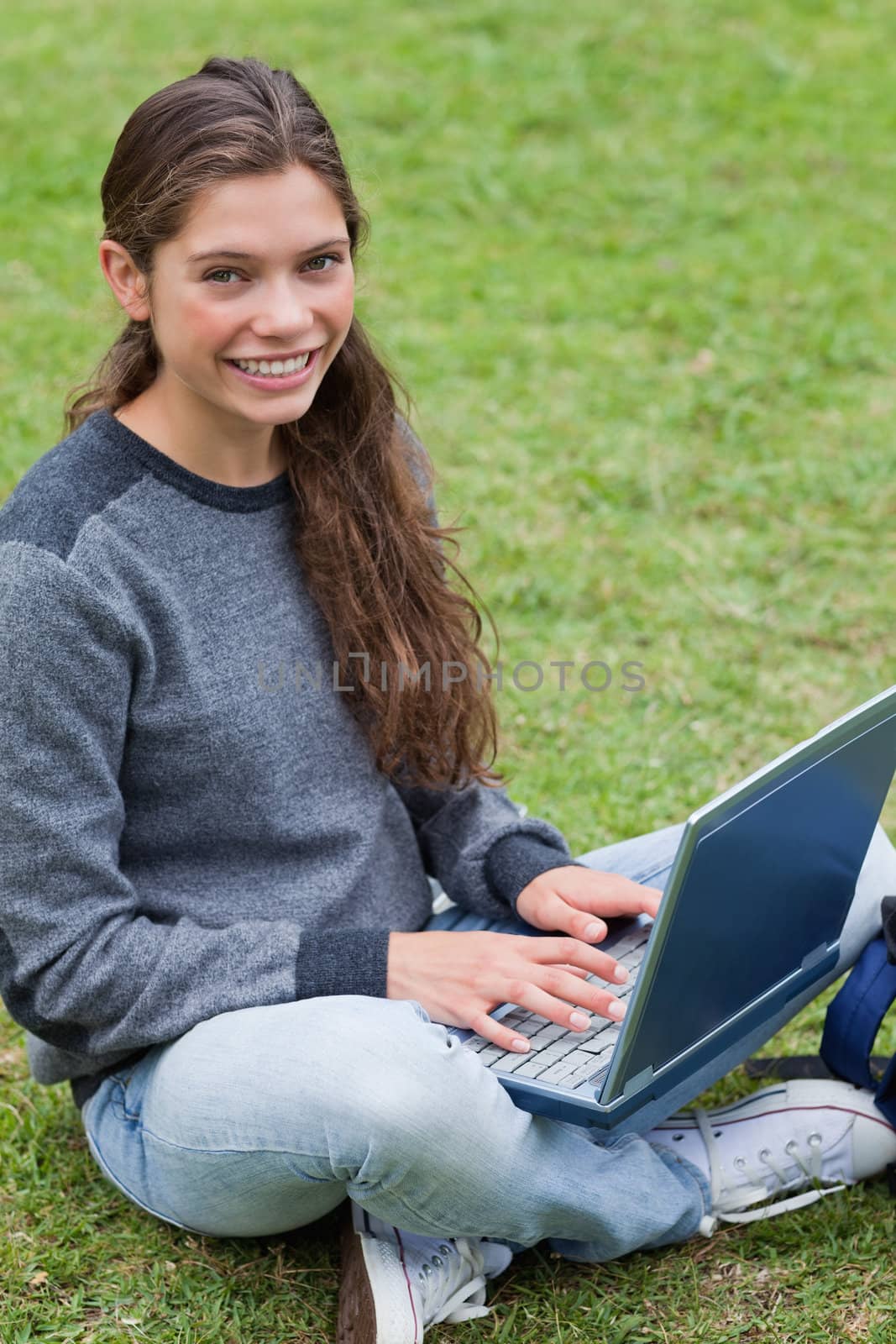 Smiling young girl sitting down on the grass in a park while typing on her laptop