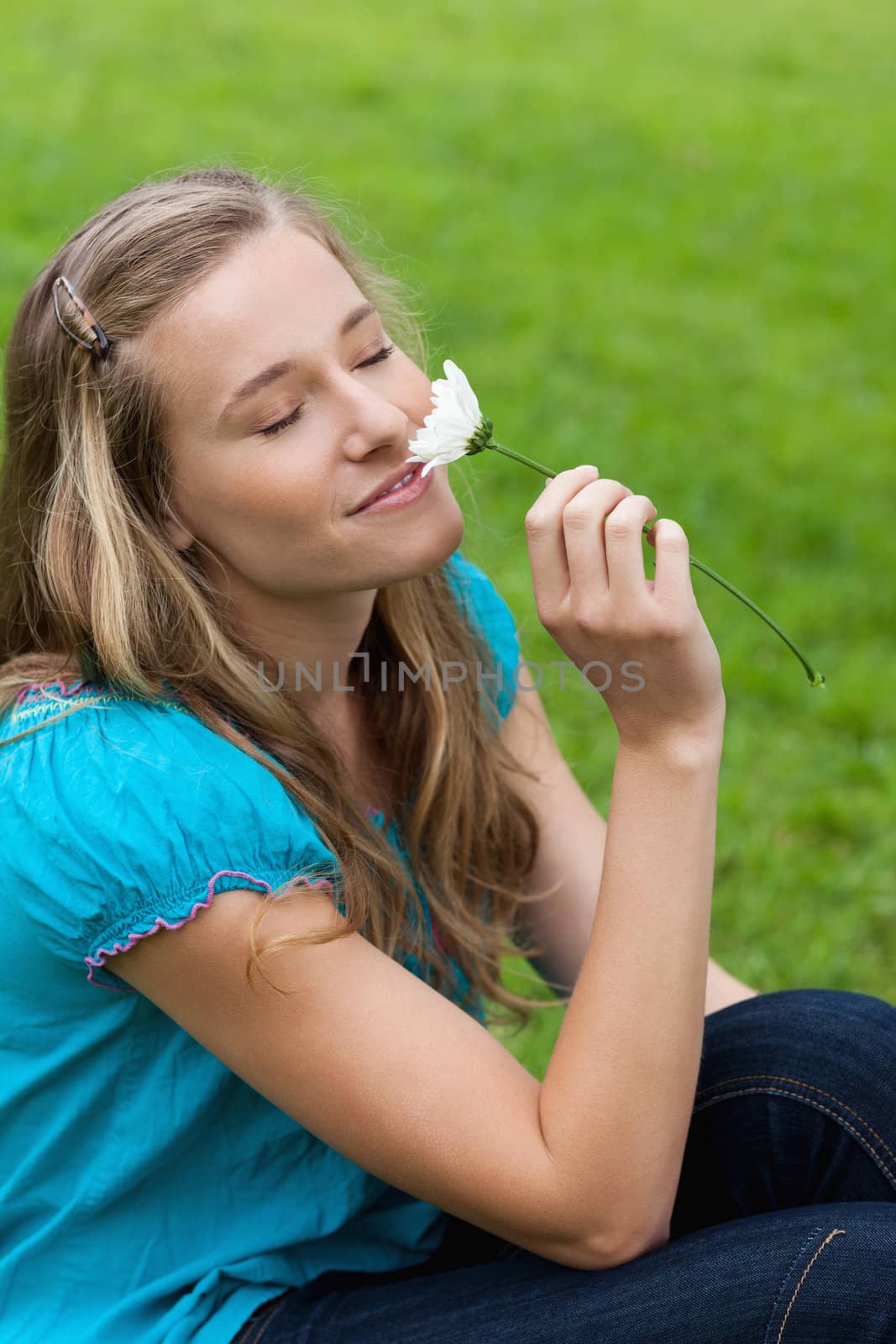 Young happy woman closing her eyes while sitting on the grass and smelling a flower