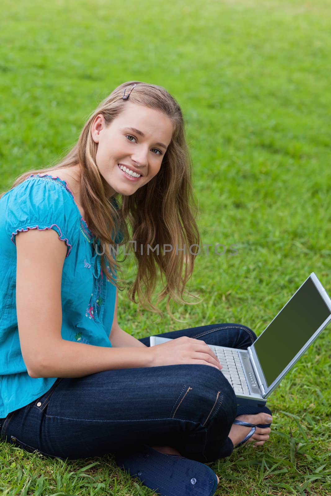 Young smiling girl sitting in a park while looking at the camera and using her laptop 