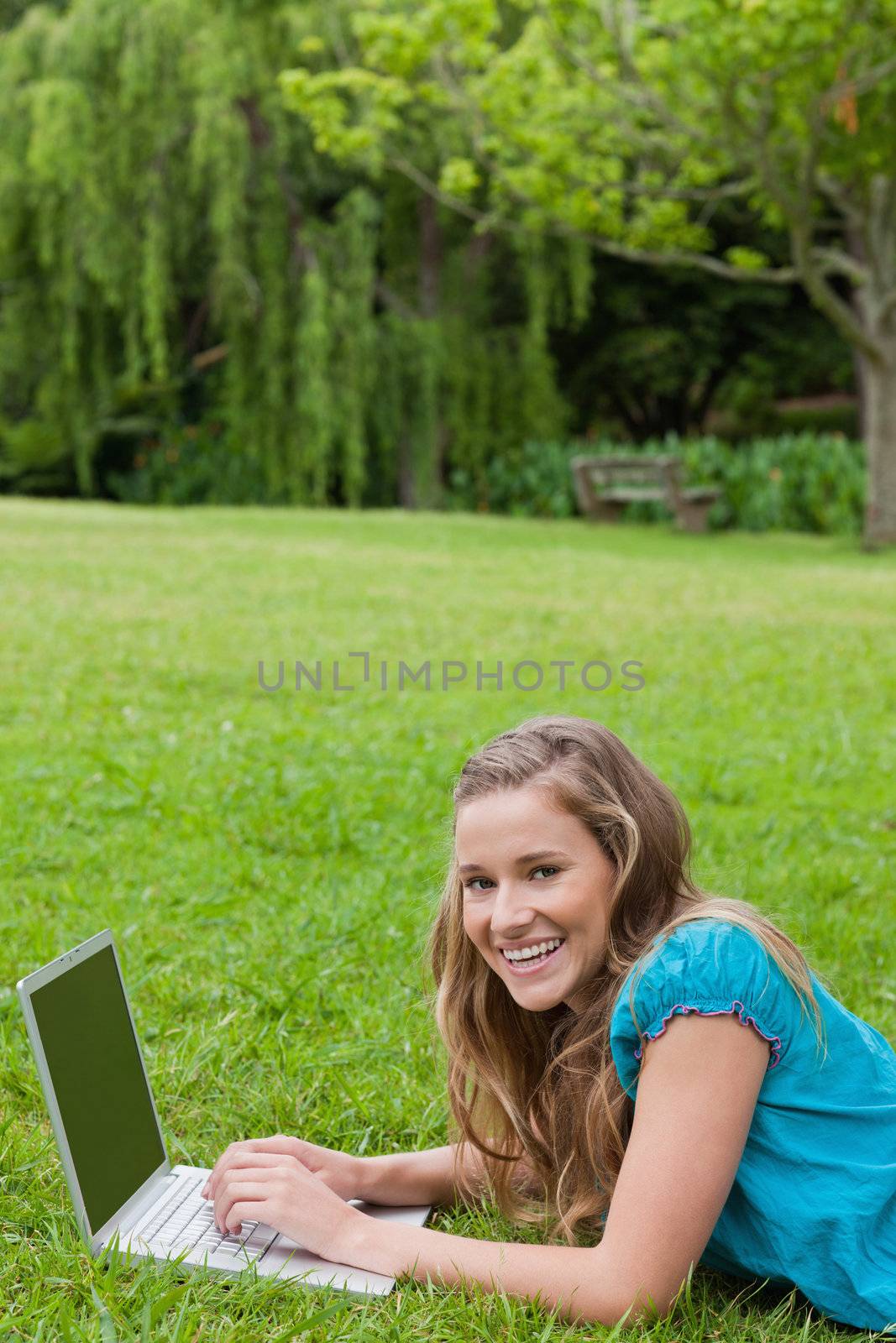 Young girl lying on the grass in a park while typing on her laptop and laughing