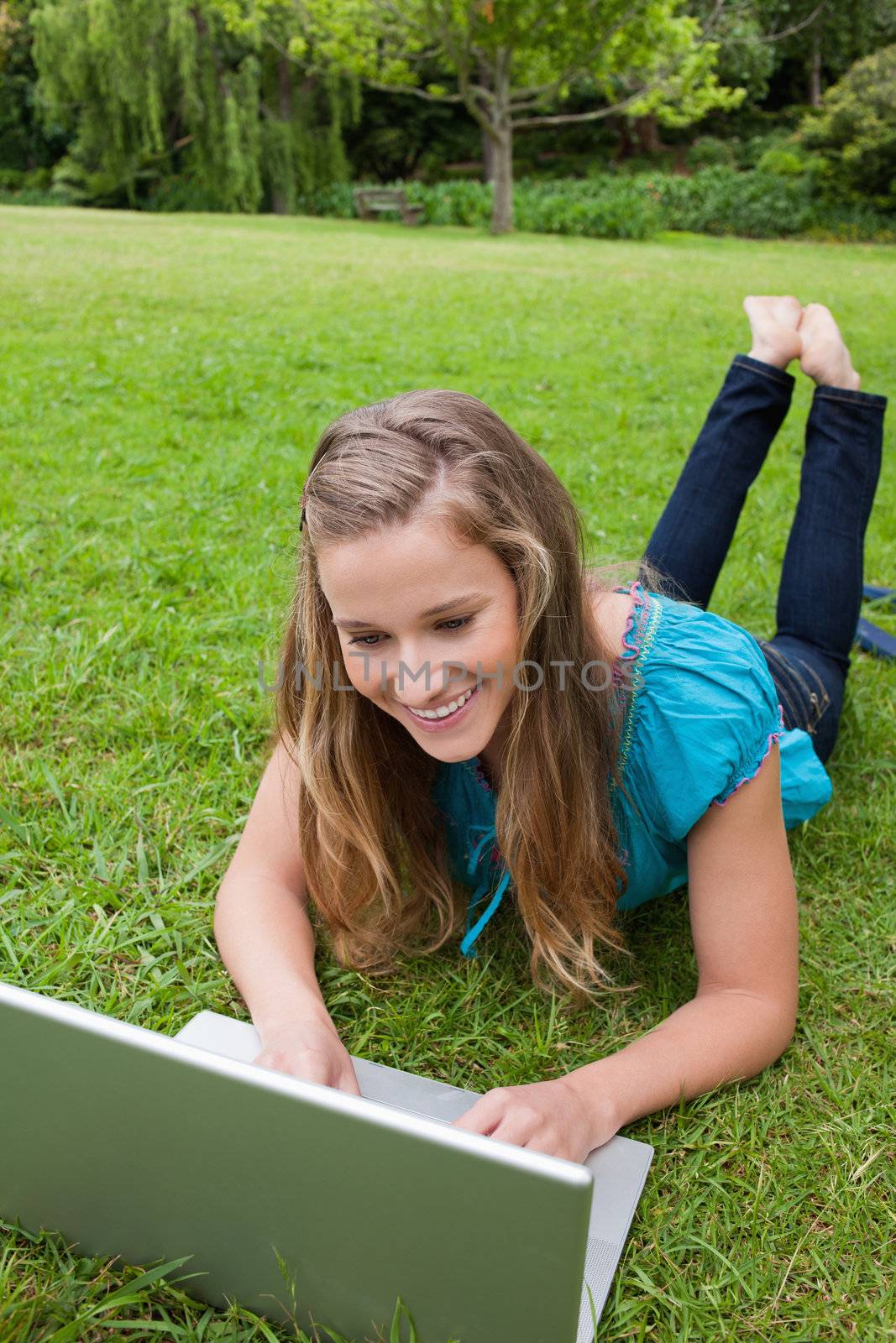 Smiling student typing on her laptop while lying down on the grass with legs raised
