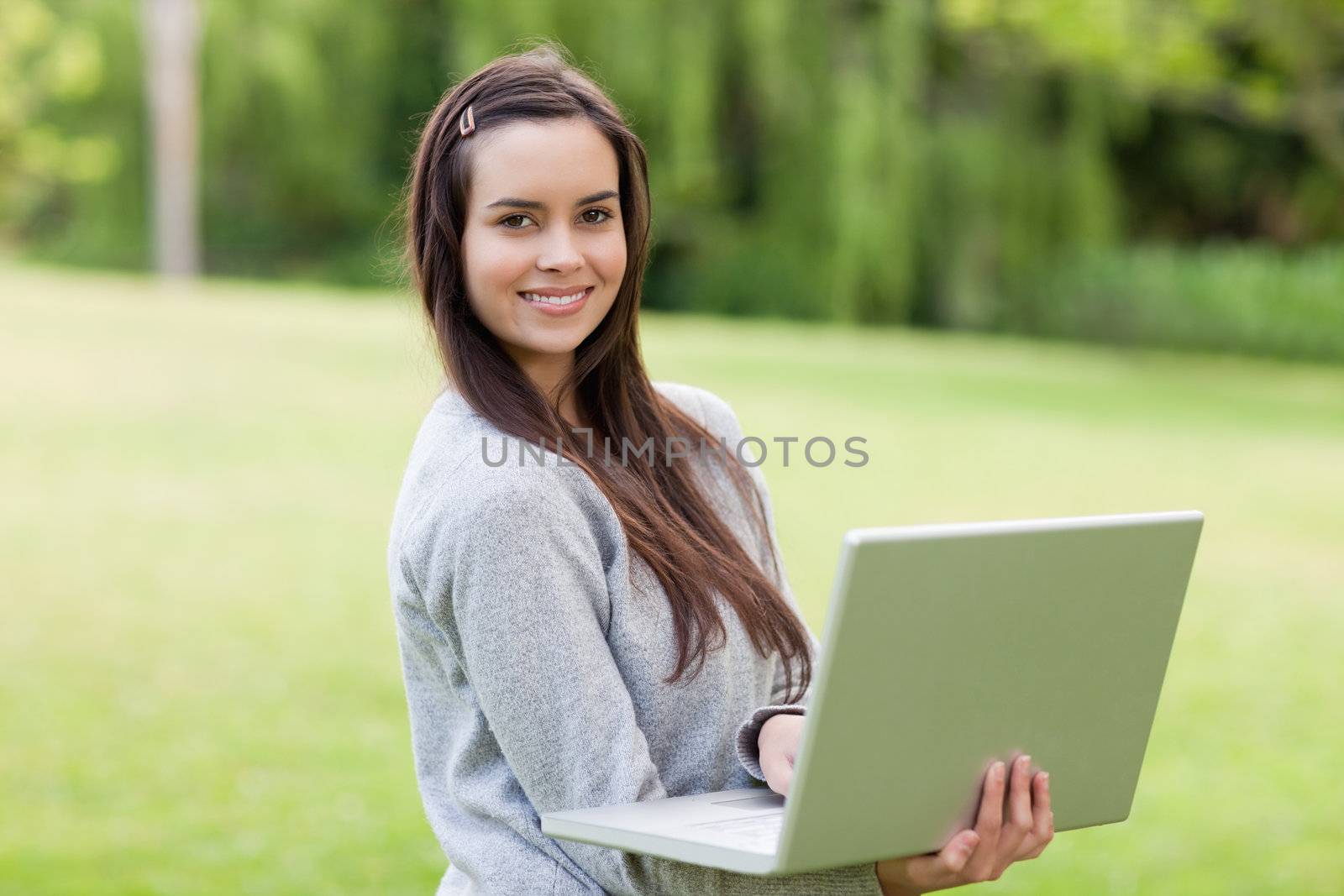 Young smiling girl standing upright in a parkland while working on her laptop
