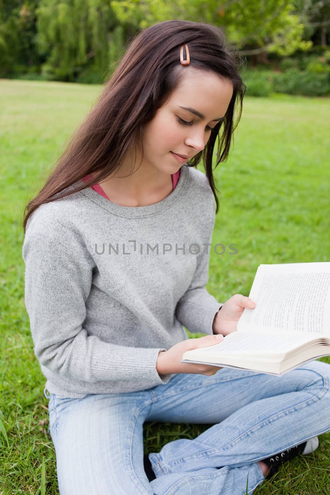 Young calm girl reading a book while sitting down on the grass in a public garden