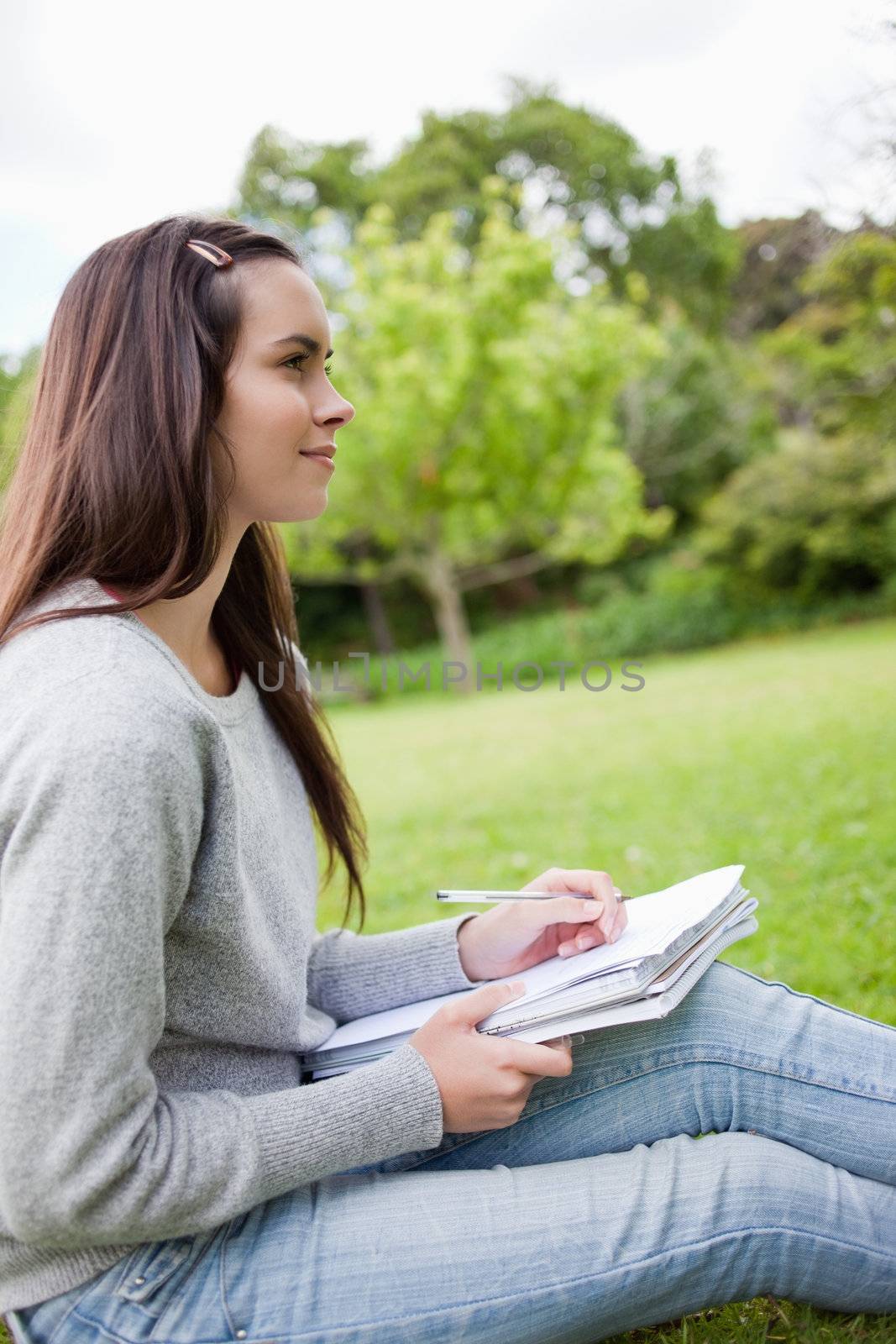 Young relaxed girl looking far away while sitting in a park with a pen and a notebook