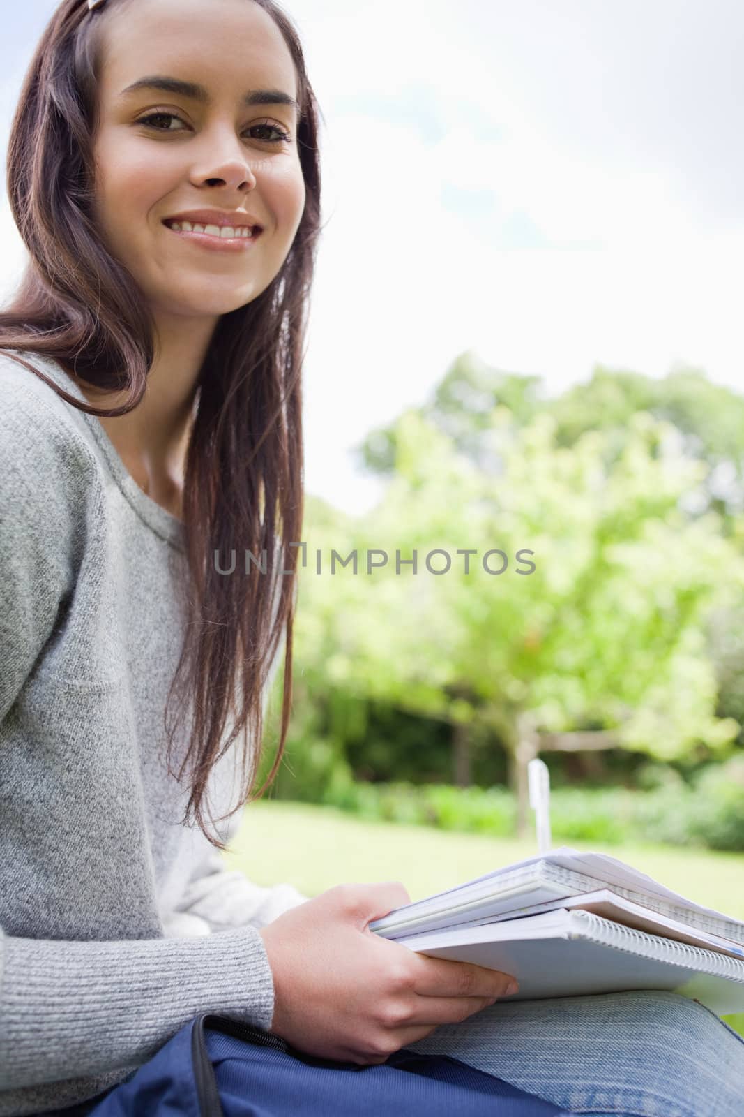 Happy student looking at the camera while sitting in a park and doing her homework