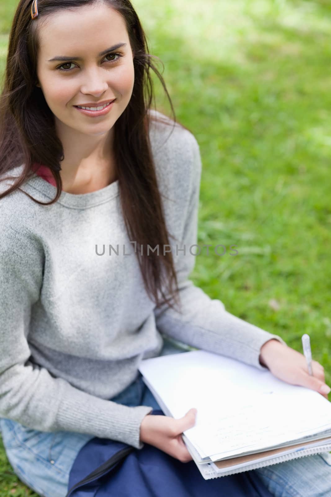 Happy student sitting in a park while looking at the camera and holding school books