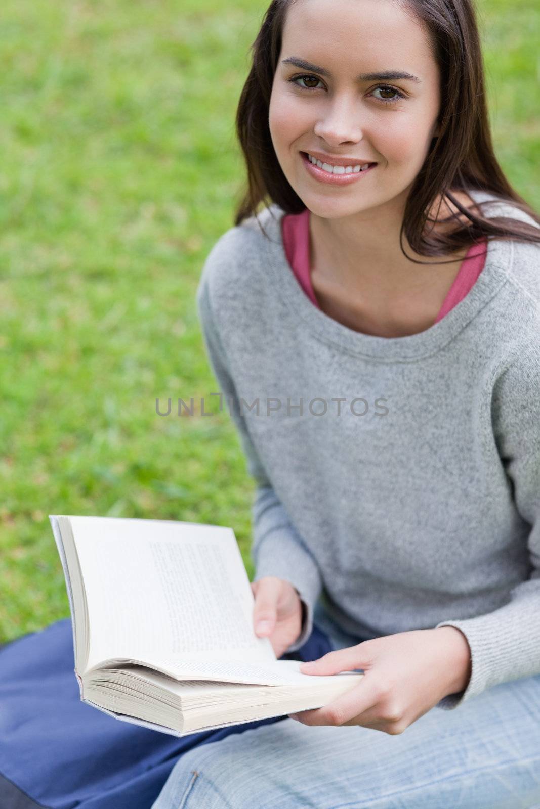 Young happy girl reading a book in a public garden while looking at the camera