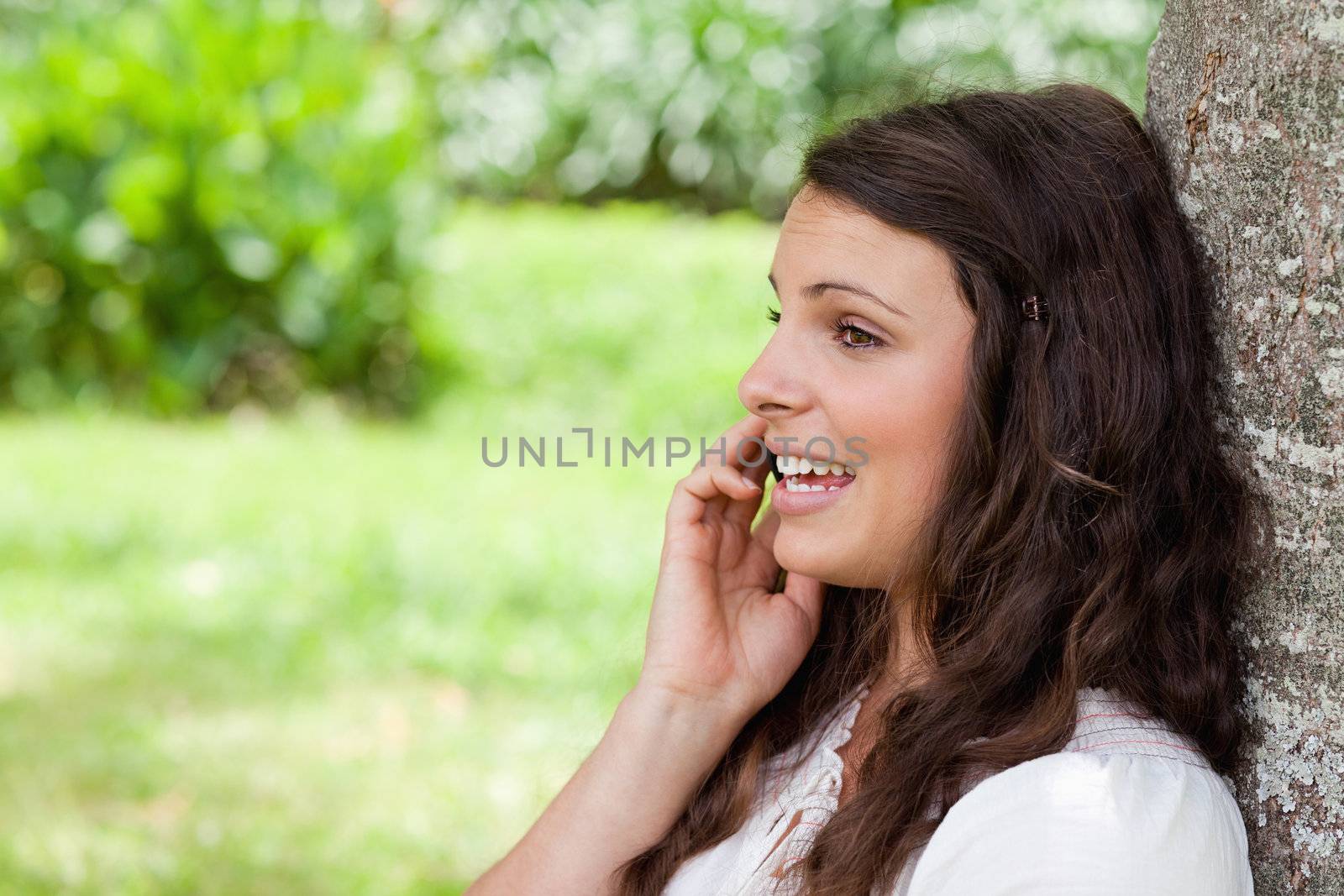 Young woman talking on the phone while leaning against a tree by Wavebreakmedia