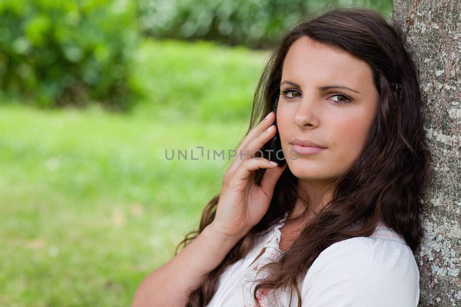 Young serious girl calling with her cellphone while sitting against a tree in a parkland